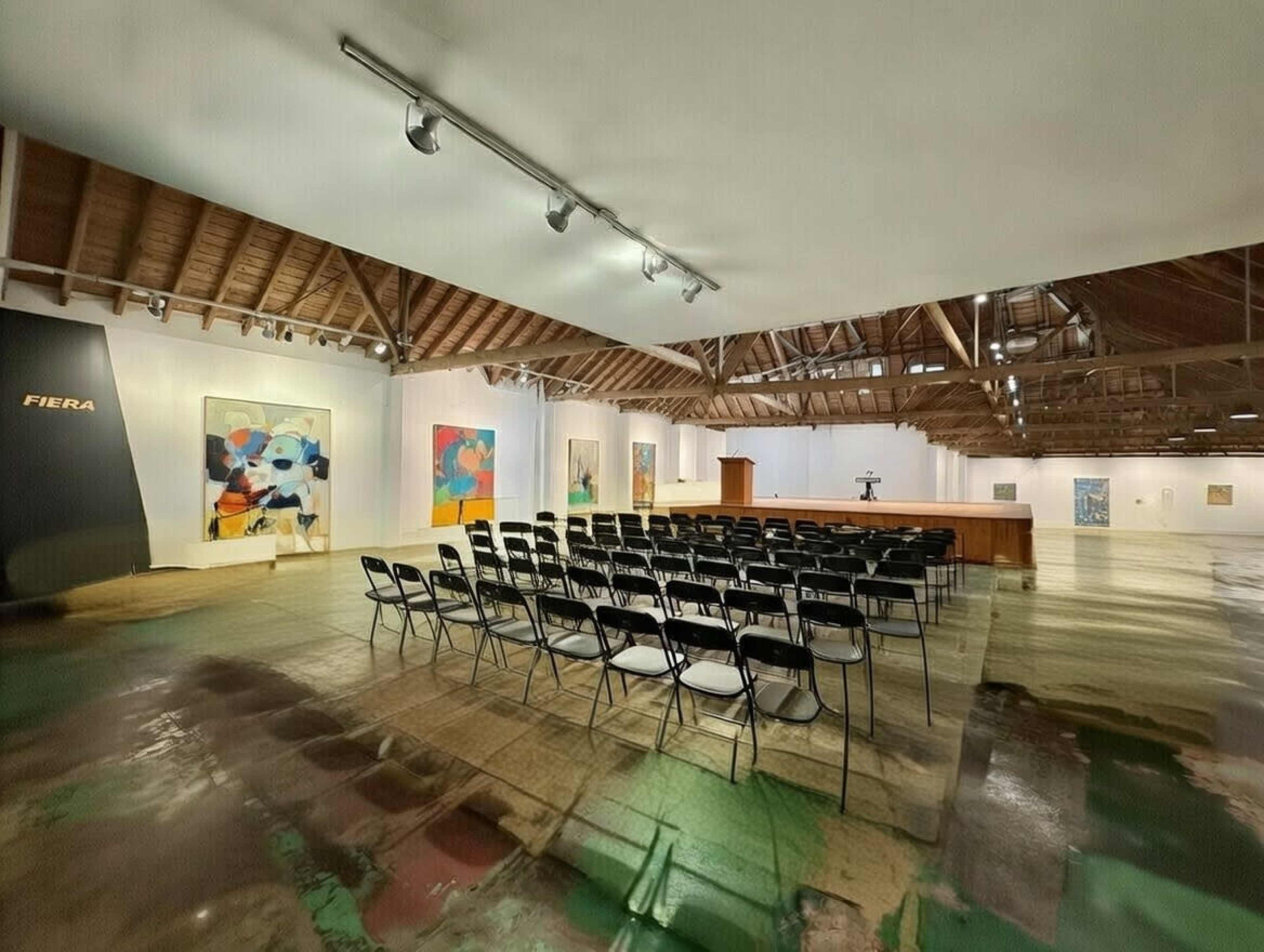 The image shows an art gallery setting with rows of black chairs arranged for an audience, colorful artworks displayed on the walls, and a podium at the front.