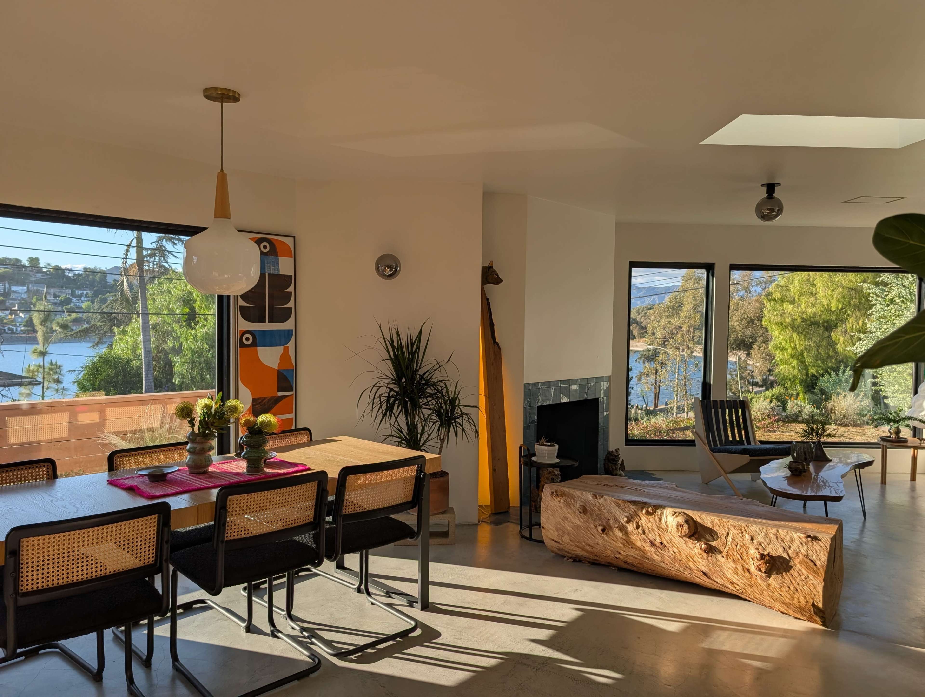 The image shows a modern dining area with a wooden table, black chairs, and large windows that overlook a scenic view, featuring natural light and minimalist decor.
