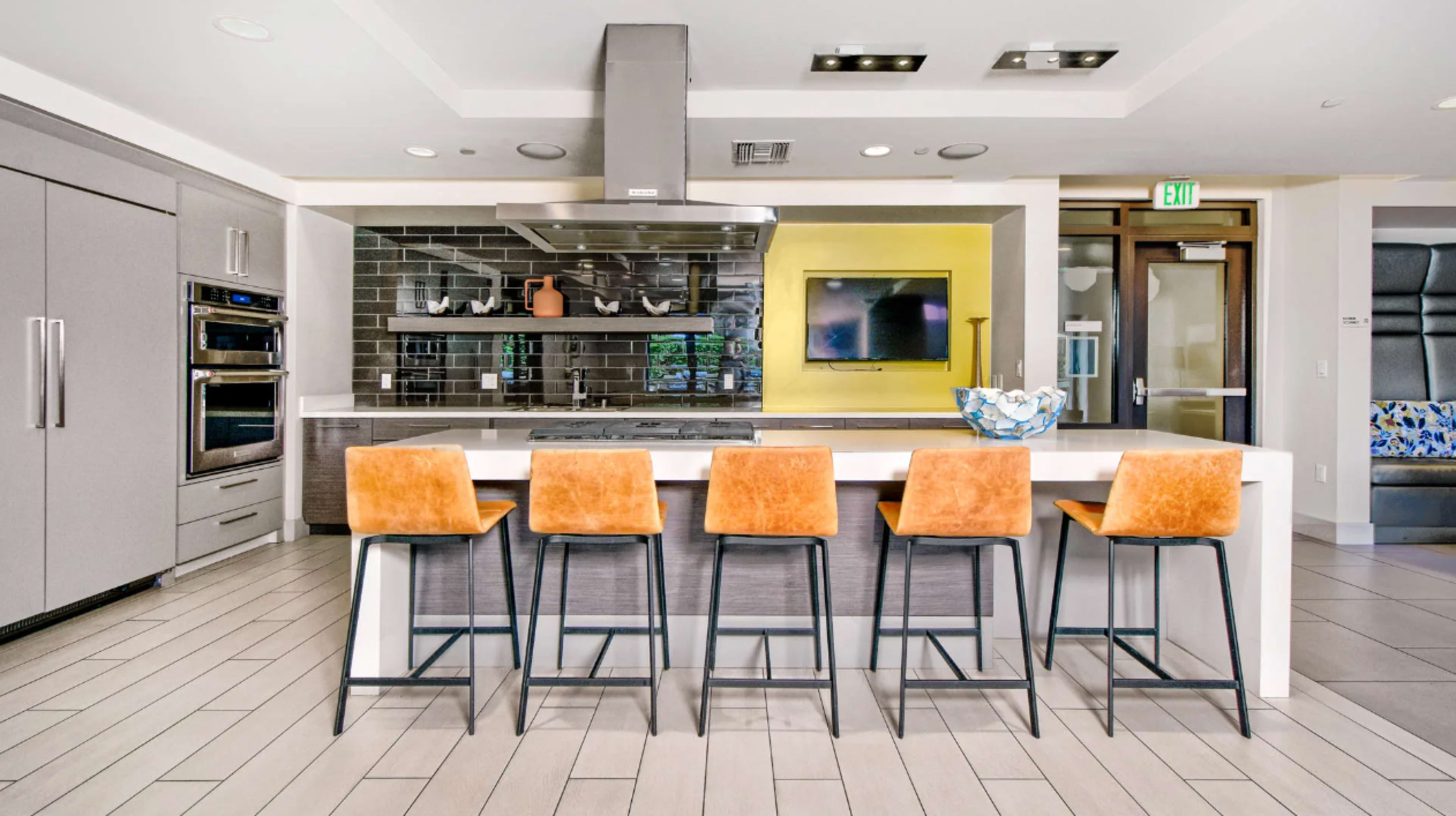 A modern kitchen features a large island with five orange bar stools and stainless steel appliances against a gray and black tiled backsplash.