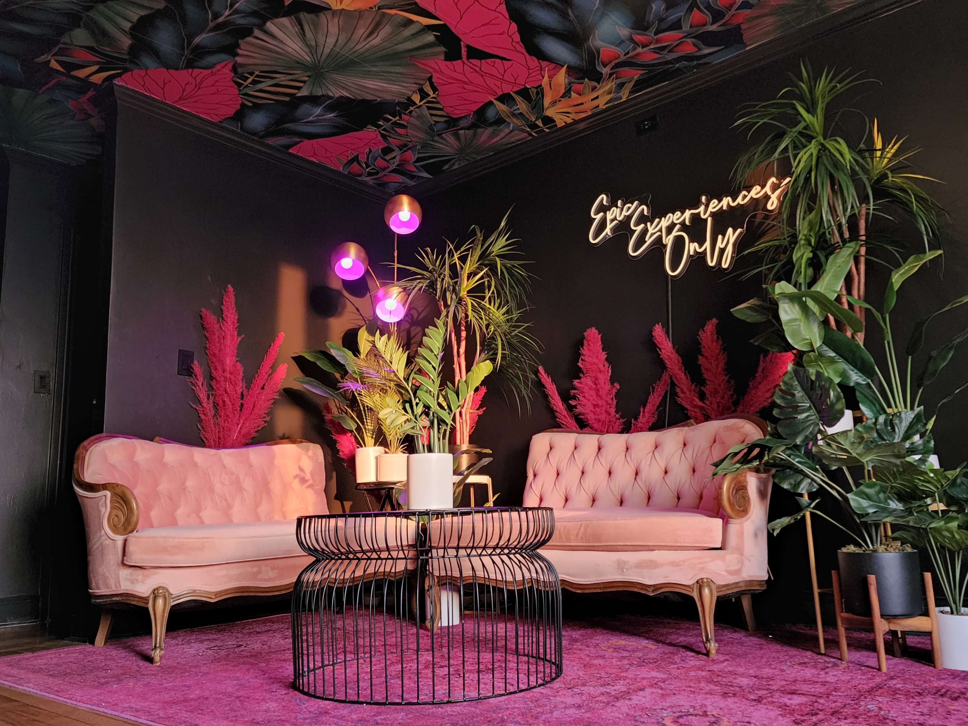 A stylish interior featuring two pink tufted sofas, a round black wire coffee table, colorful plants, and a neon sign that reads "Epic Experiences Only" against a dark wall.
