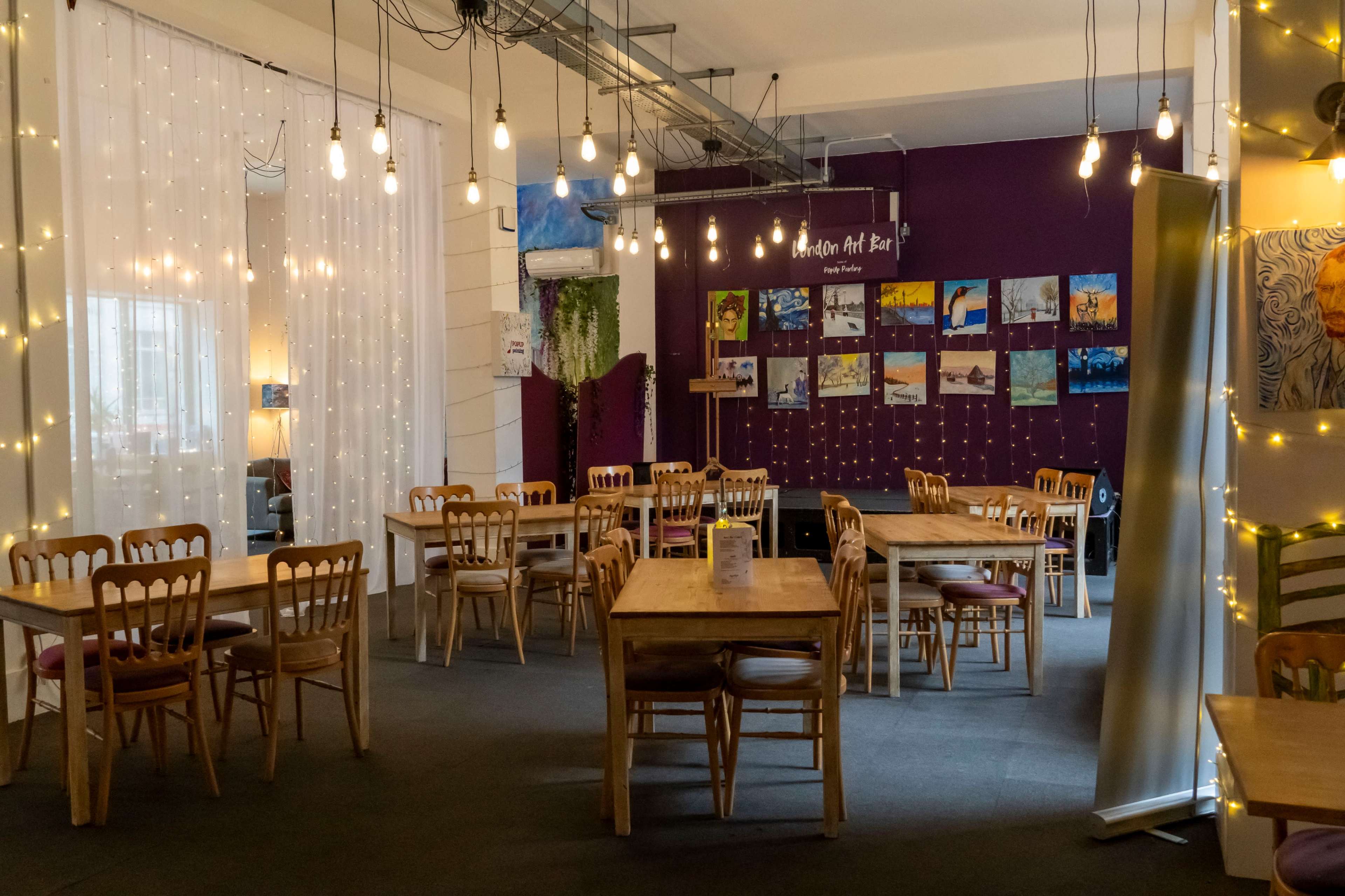 The image shows a well-lit café or art bar featuring multiple wooden tables and chairs arranged in a spacious area with string lights and artwork on the walls.