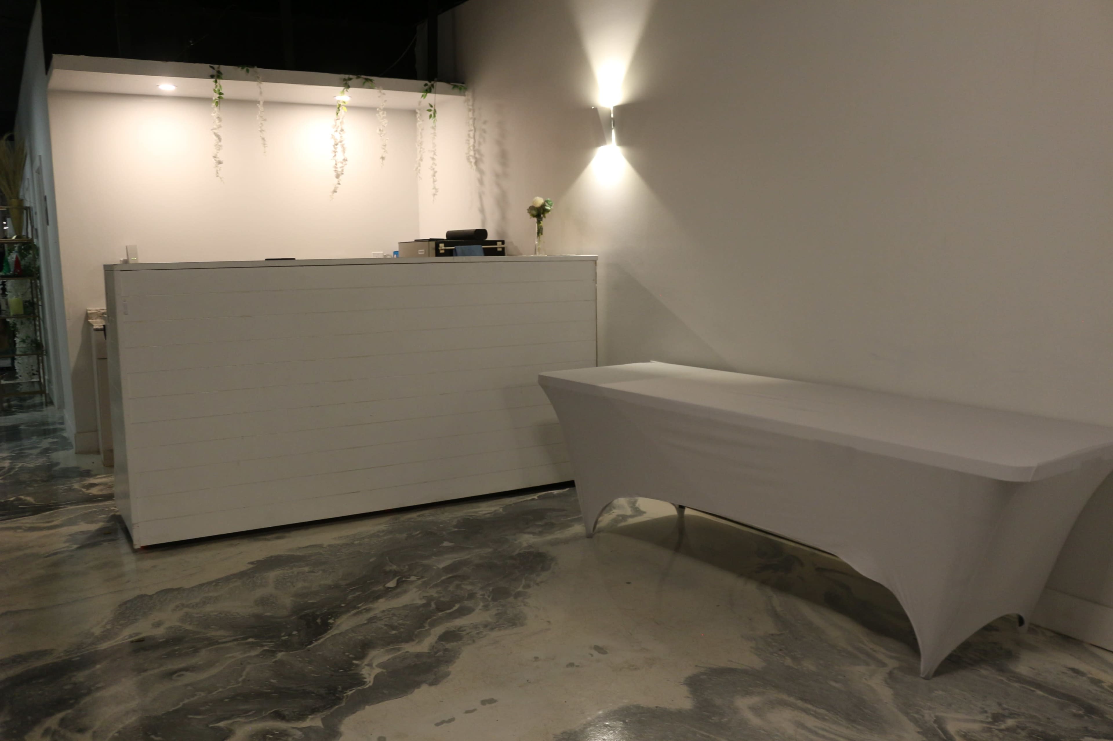 The image shows a minimalist reception area with a white front desk and a covered table against a plain wall.