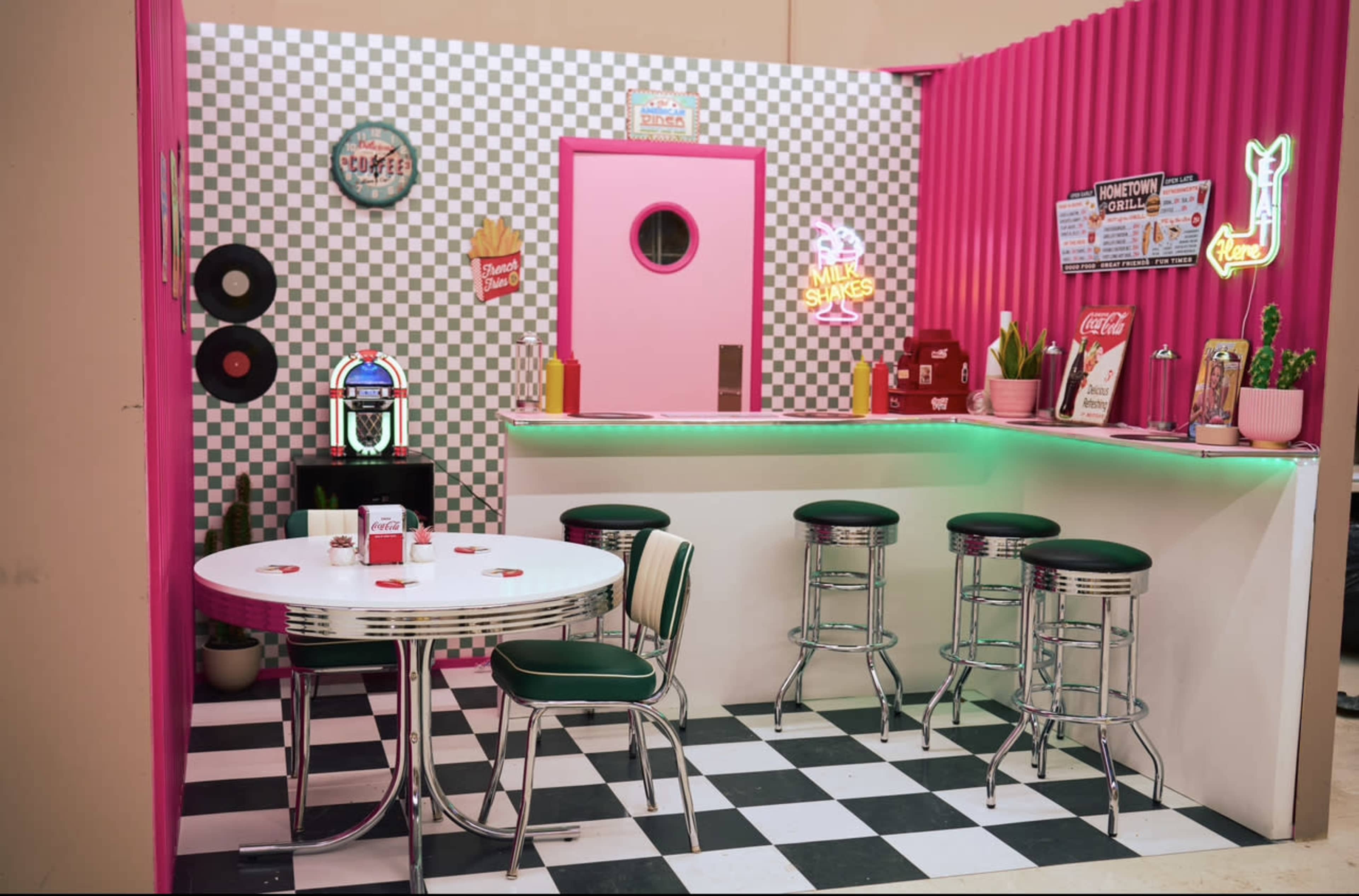 The image shows a retro diner setup featuring a pink and black checkered theme, with a round table and chairs, a bar counter, and neon signs.
