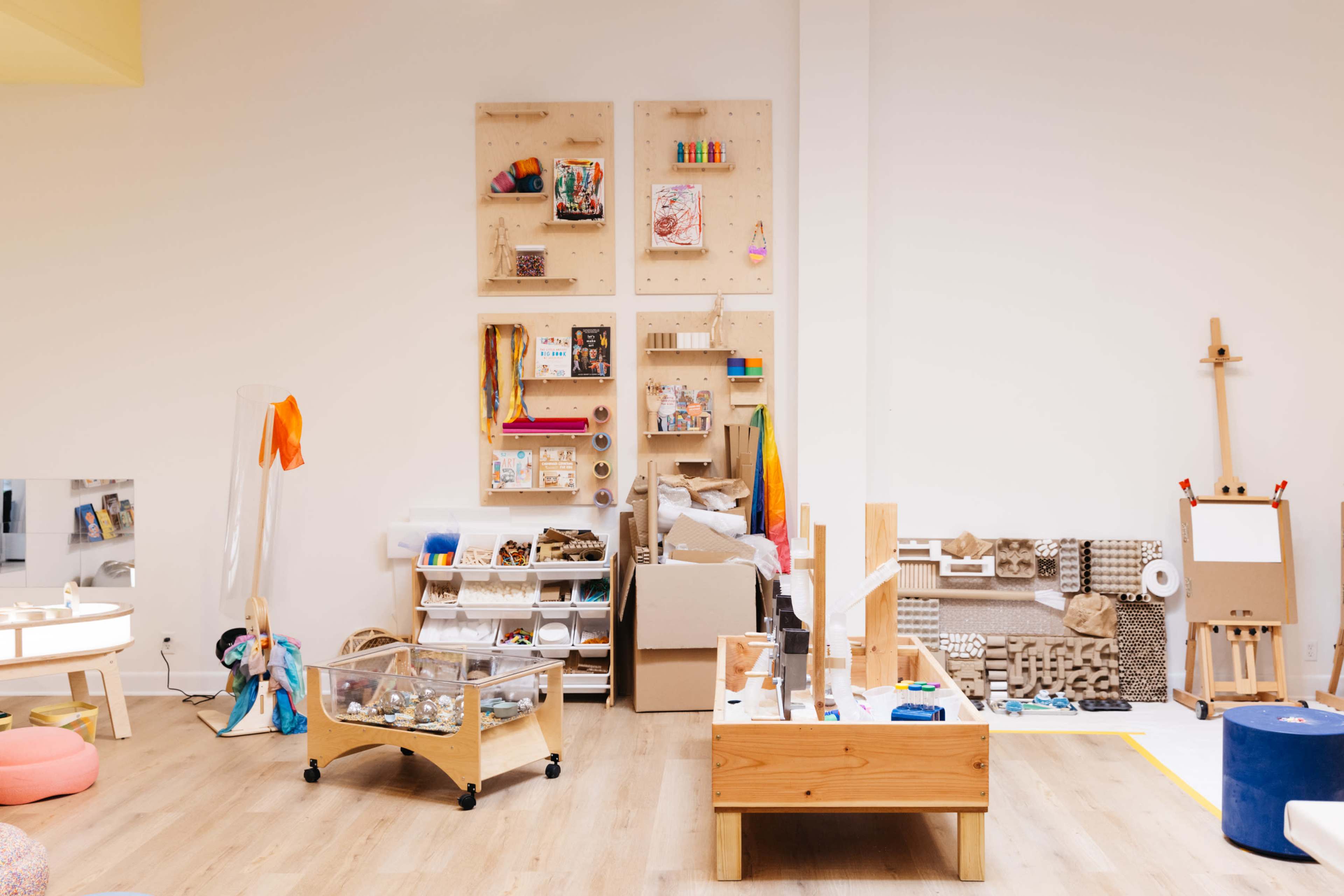 The image shows a brightly lit classroom or art studio featuring various workstations, storage units filled with craft supplies, an easel, and a textured wall display.
