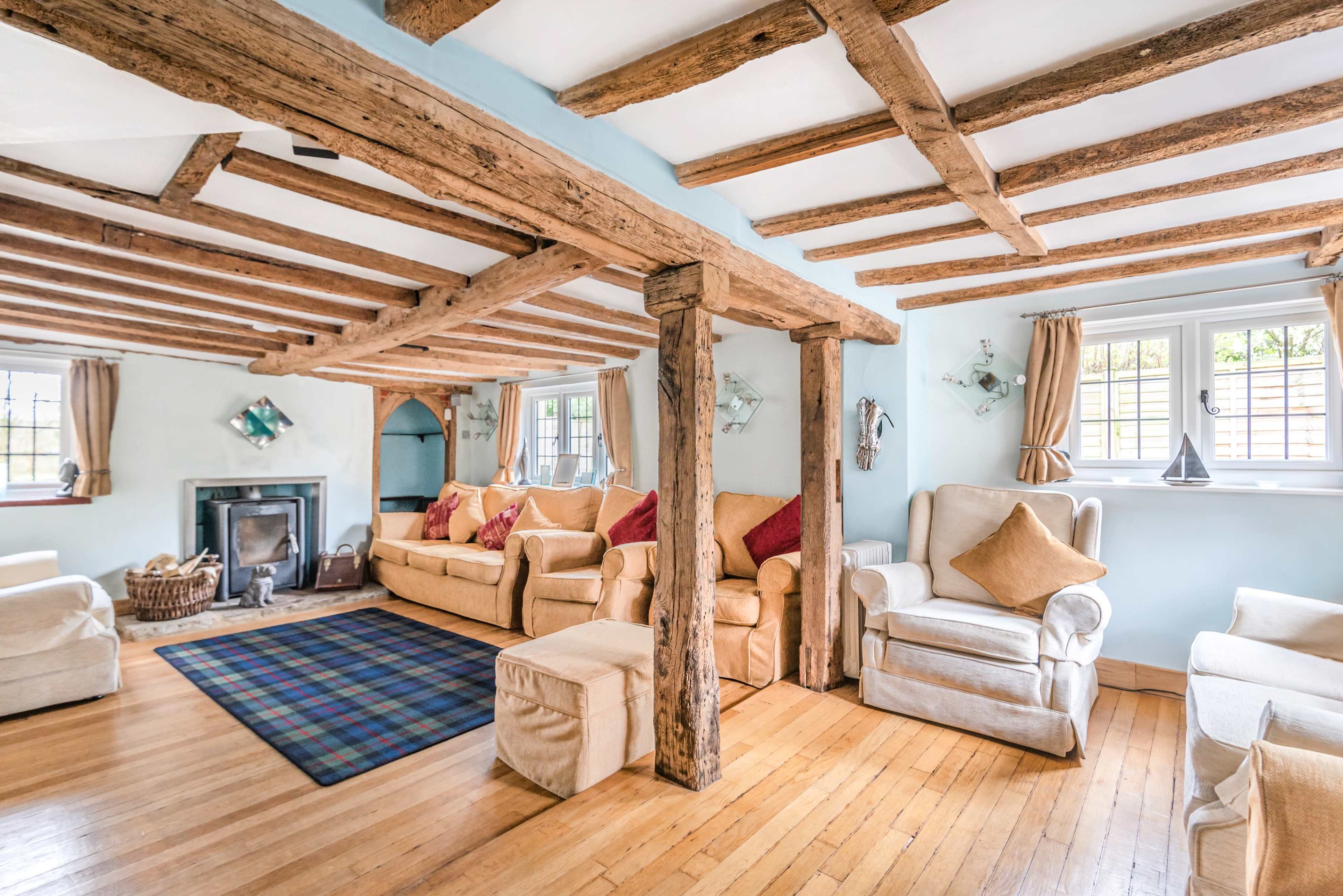 The living room features exposed wooden beams, beige upholstered sofas, a blue and black plaid rug, and a fireplace, with large windows allowing natural light to enter.