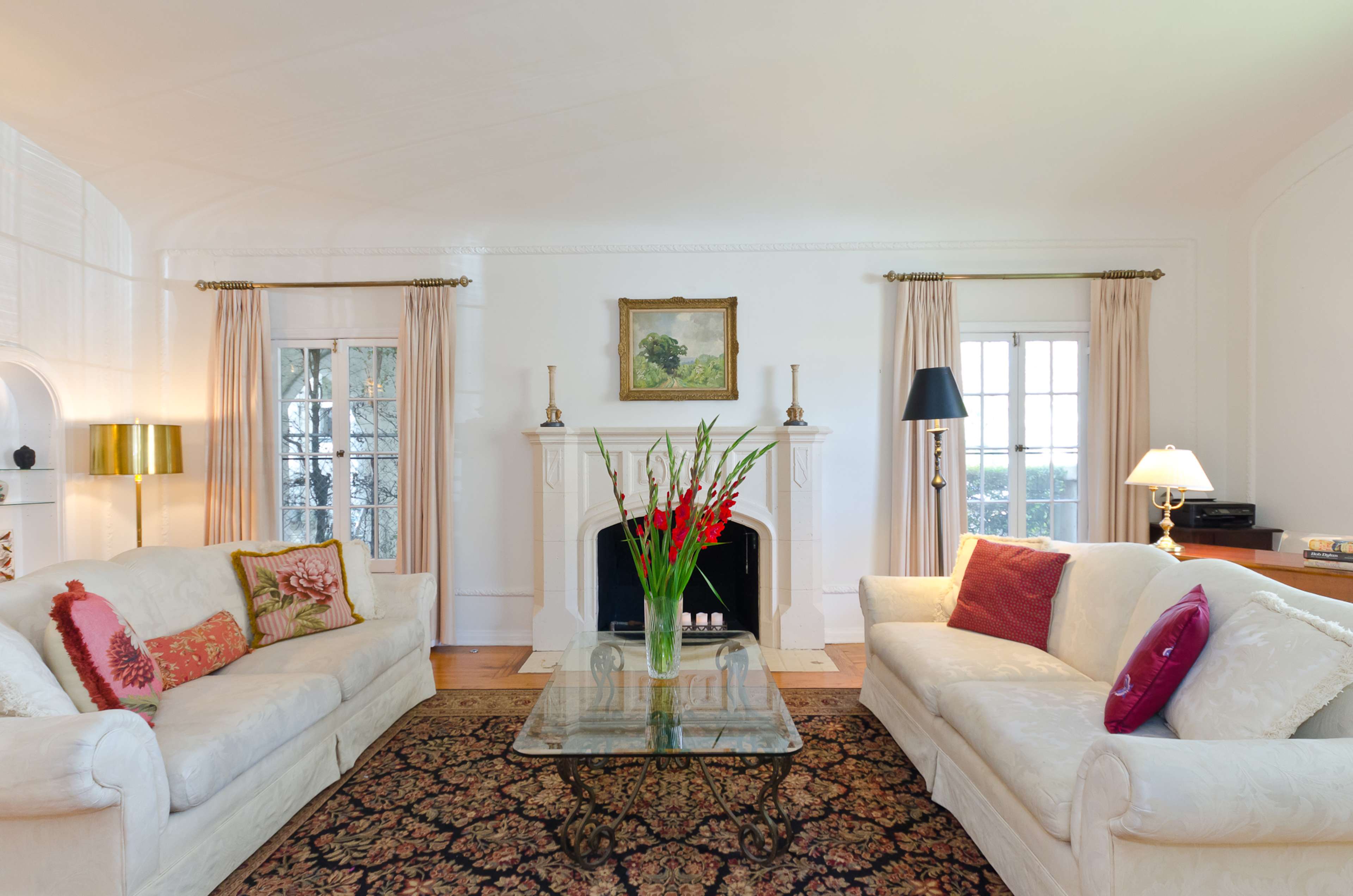 A bright living room features two white sofas with decorative pillows, a glass coffee table, and a fireplace, framed by large windows and elegant curtains.