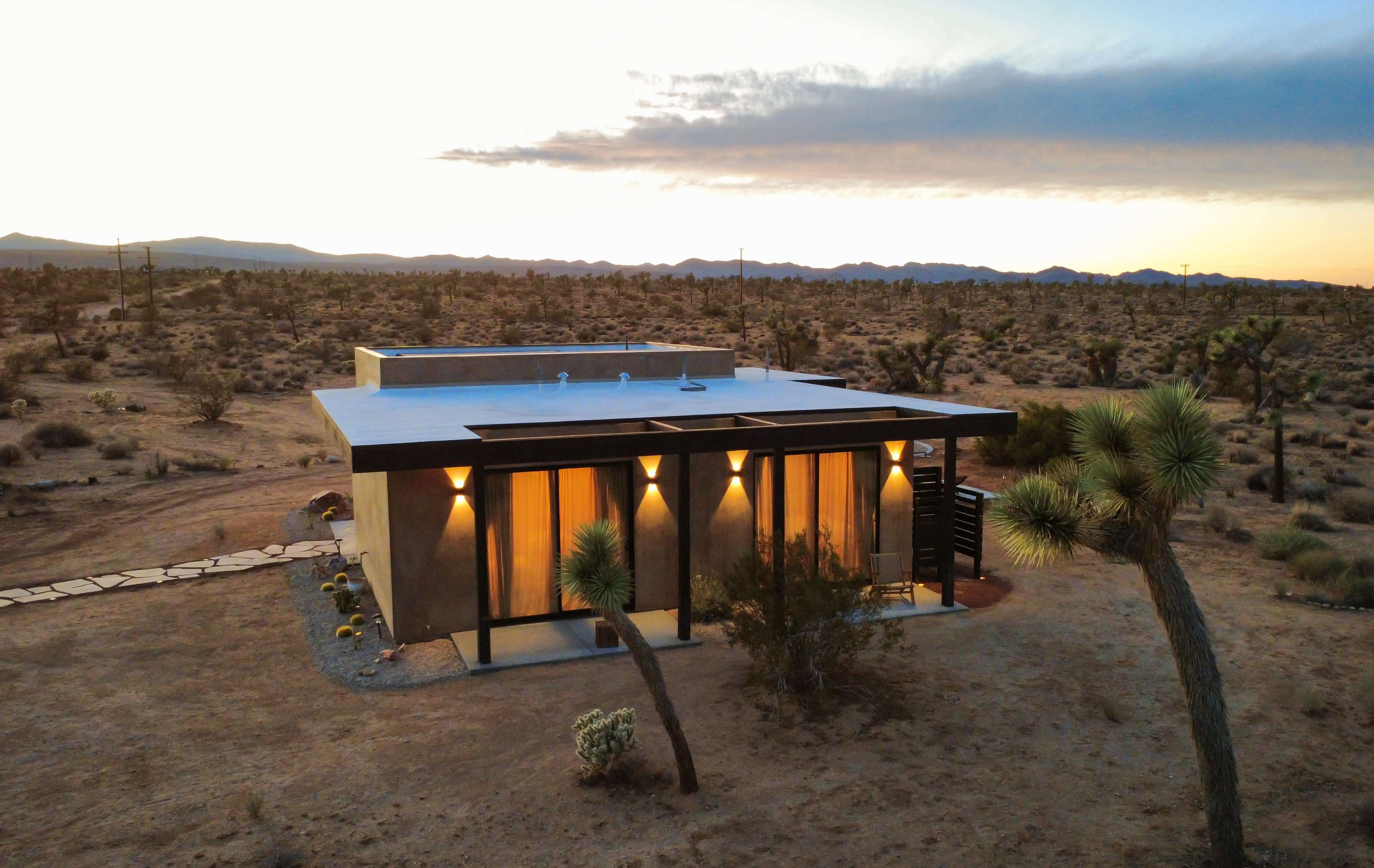 A modern house with large windows is situated in a desert landscape at dusk, surrounded by sparse vegetation and mountains in the background.