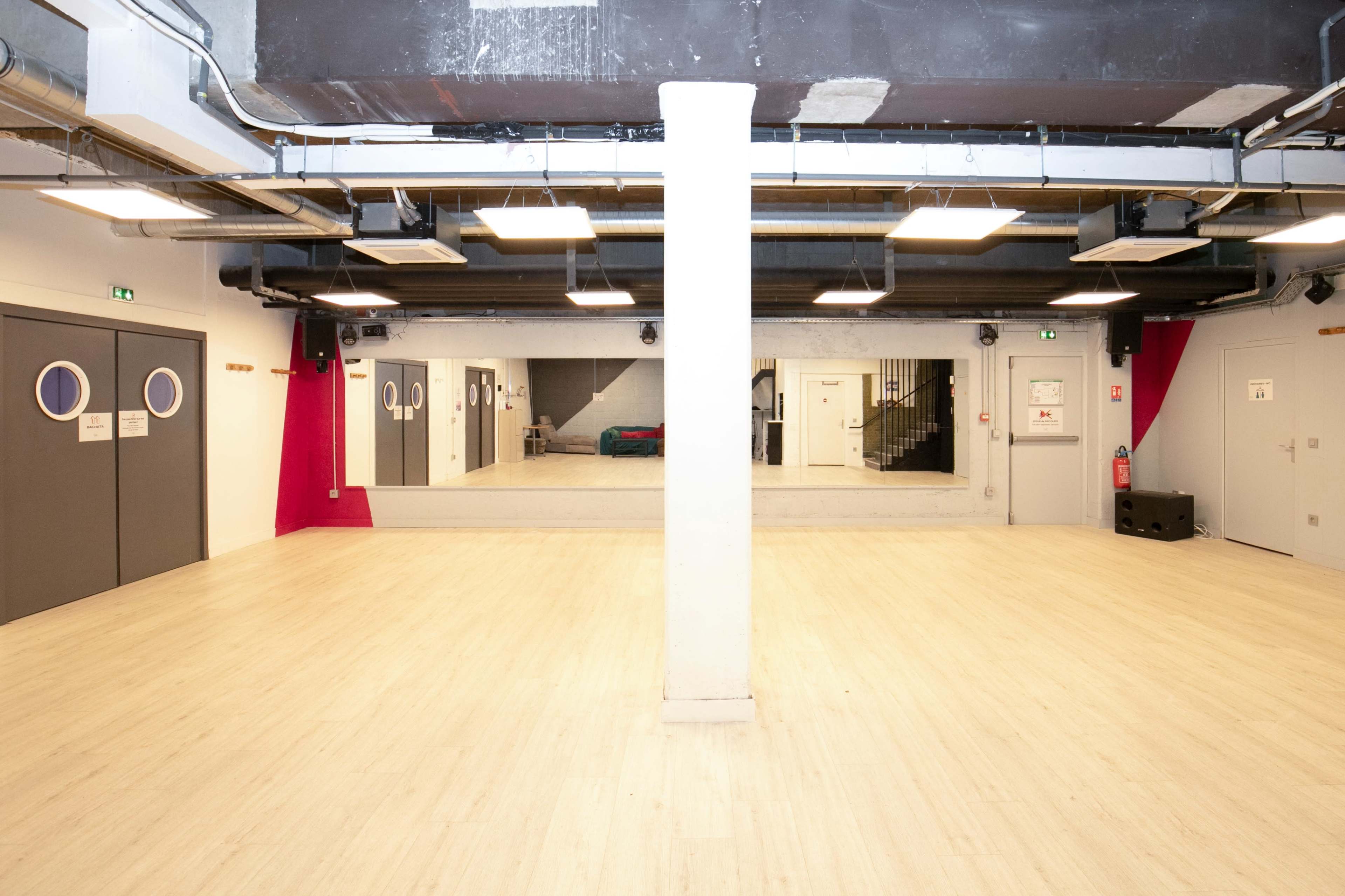 A spacious, empty room with wooden flooring and mirrored walls, featuring a central column and several doors.