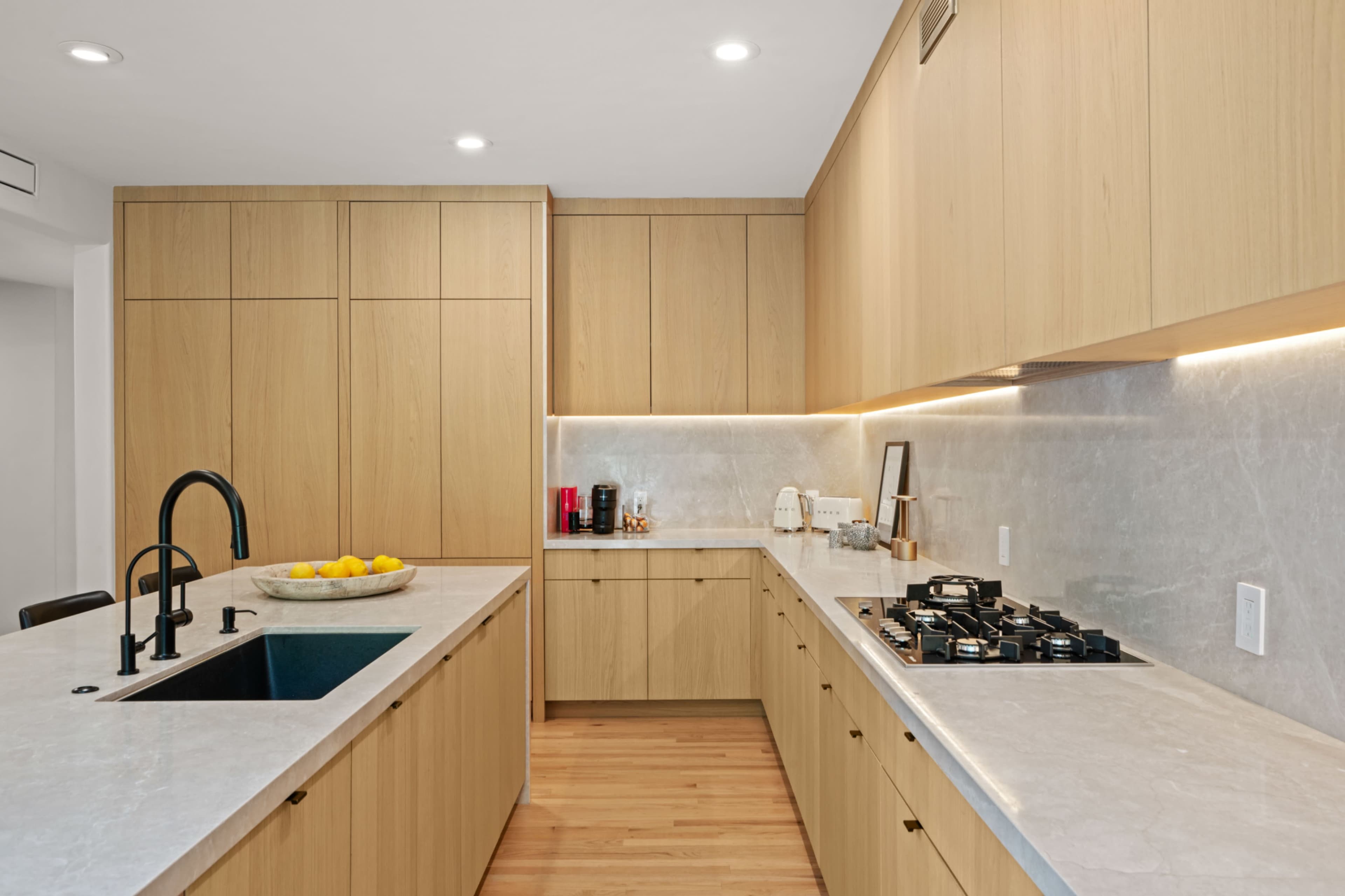 The image depicts a modern kitchen featuring light wood cabinetry, a marble countertop, an undermount sink, and a gas stove.
