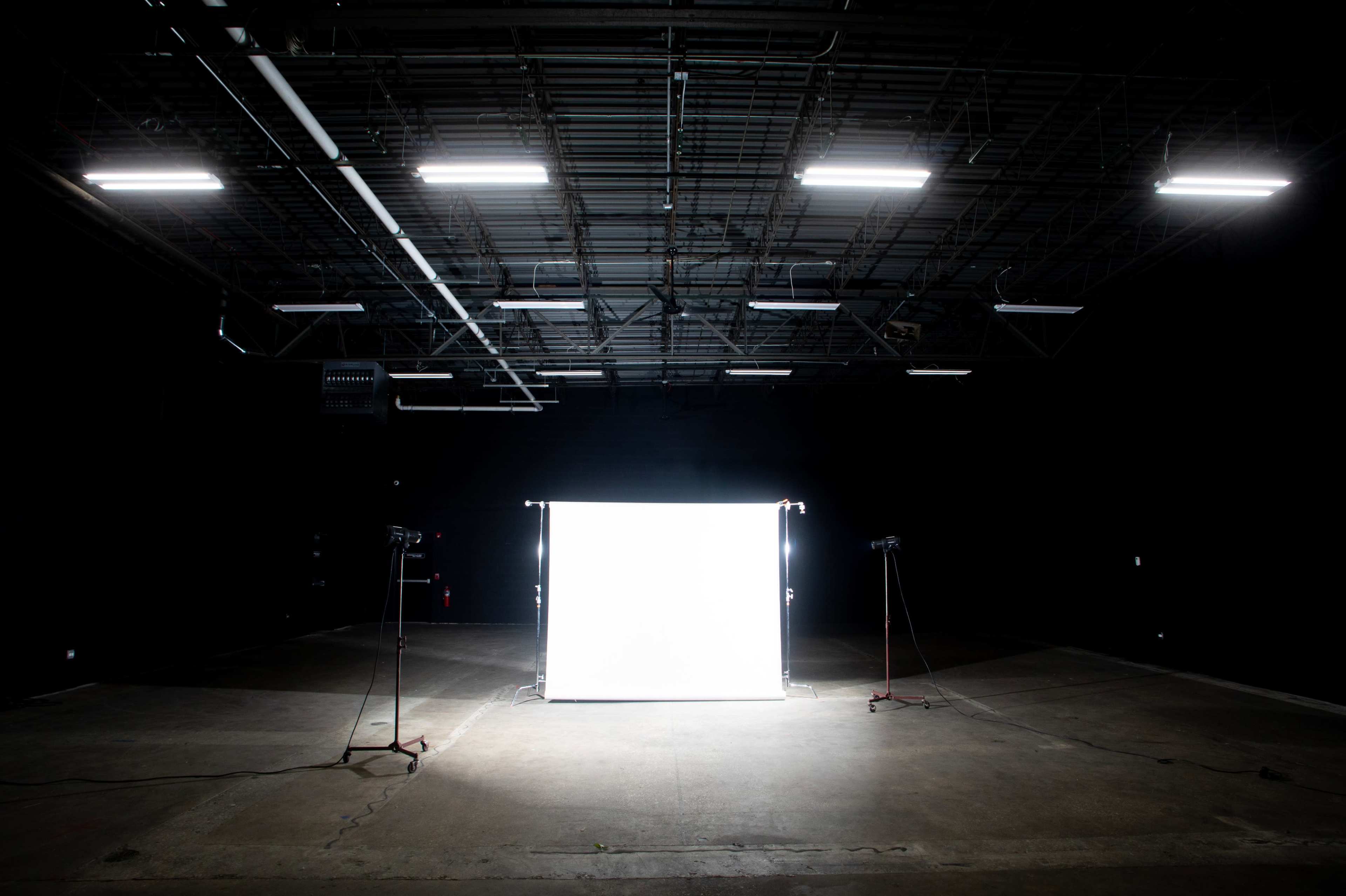 The image shows a dimly lit warehouse space with a bright white backdrop illuminated by two lights on stands.