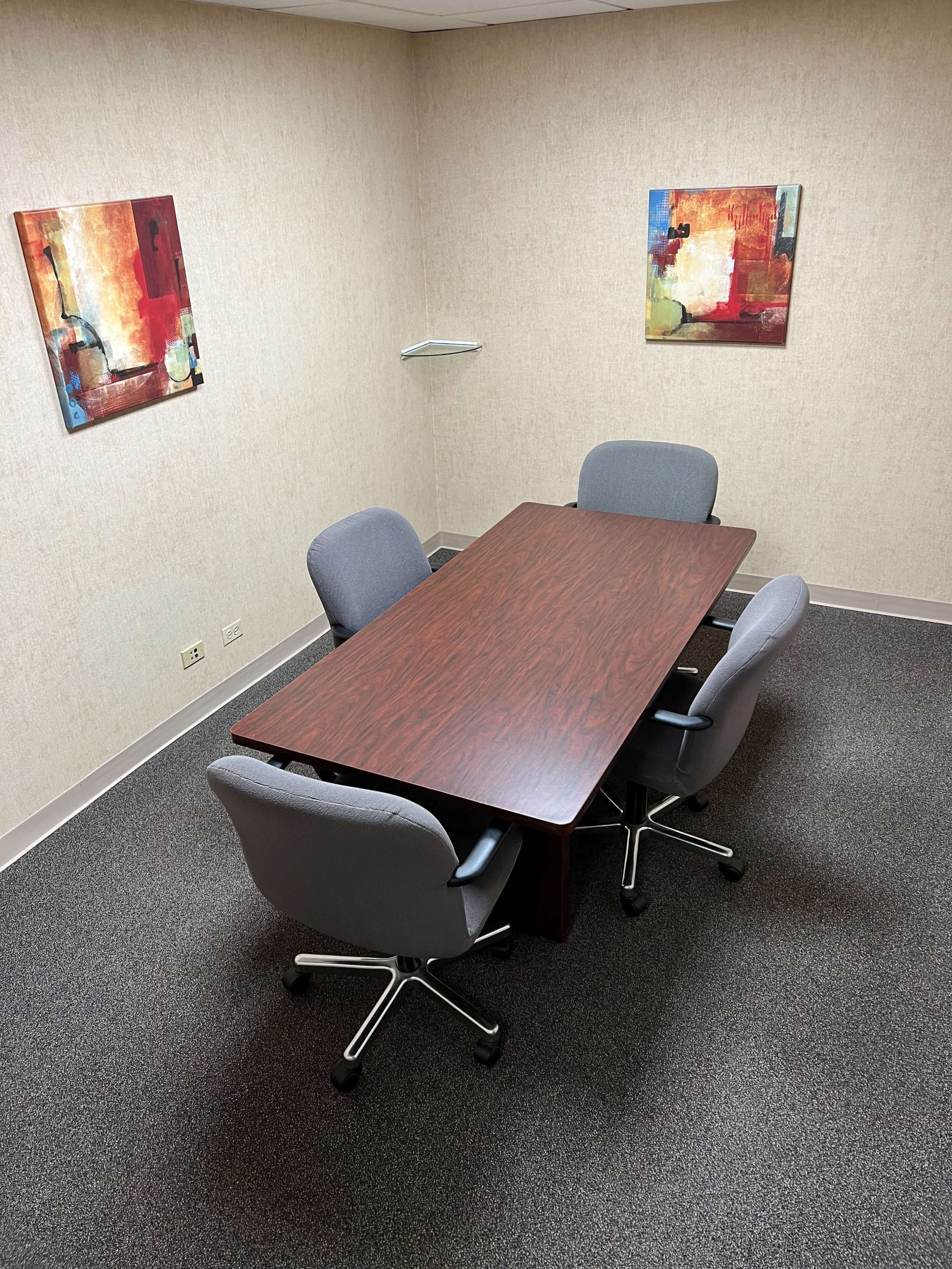 A conference room features a rectangular table surrounded by four gray chairs and two abstract paintings on the walls.
