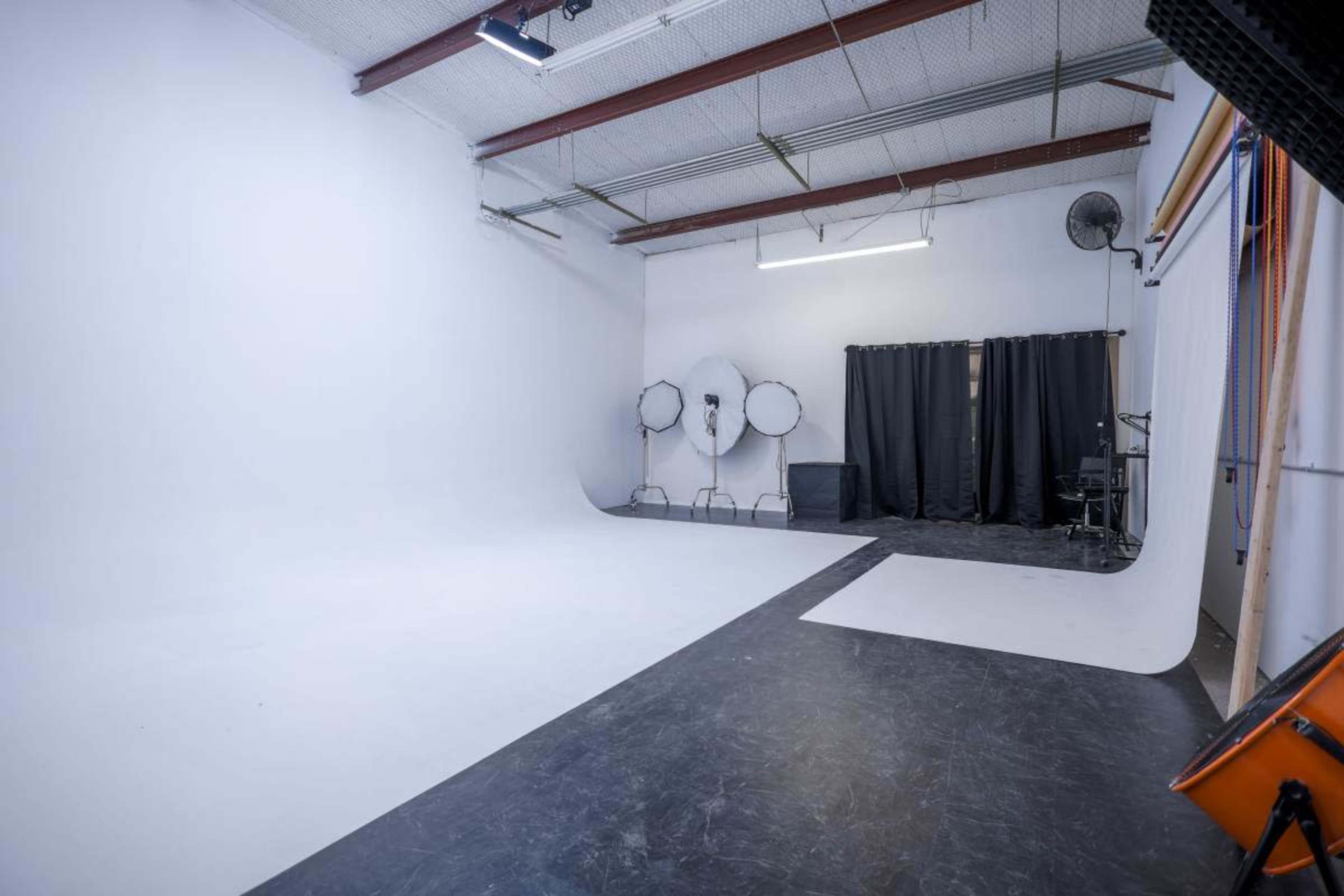 The image shows a photography studio with a white backdrop, a black floor, and softbox lighting equipment positioned at the rear.