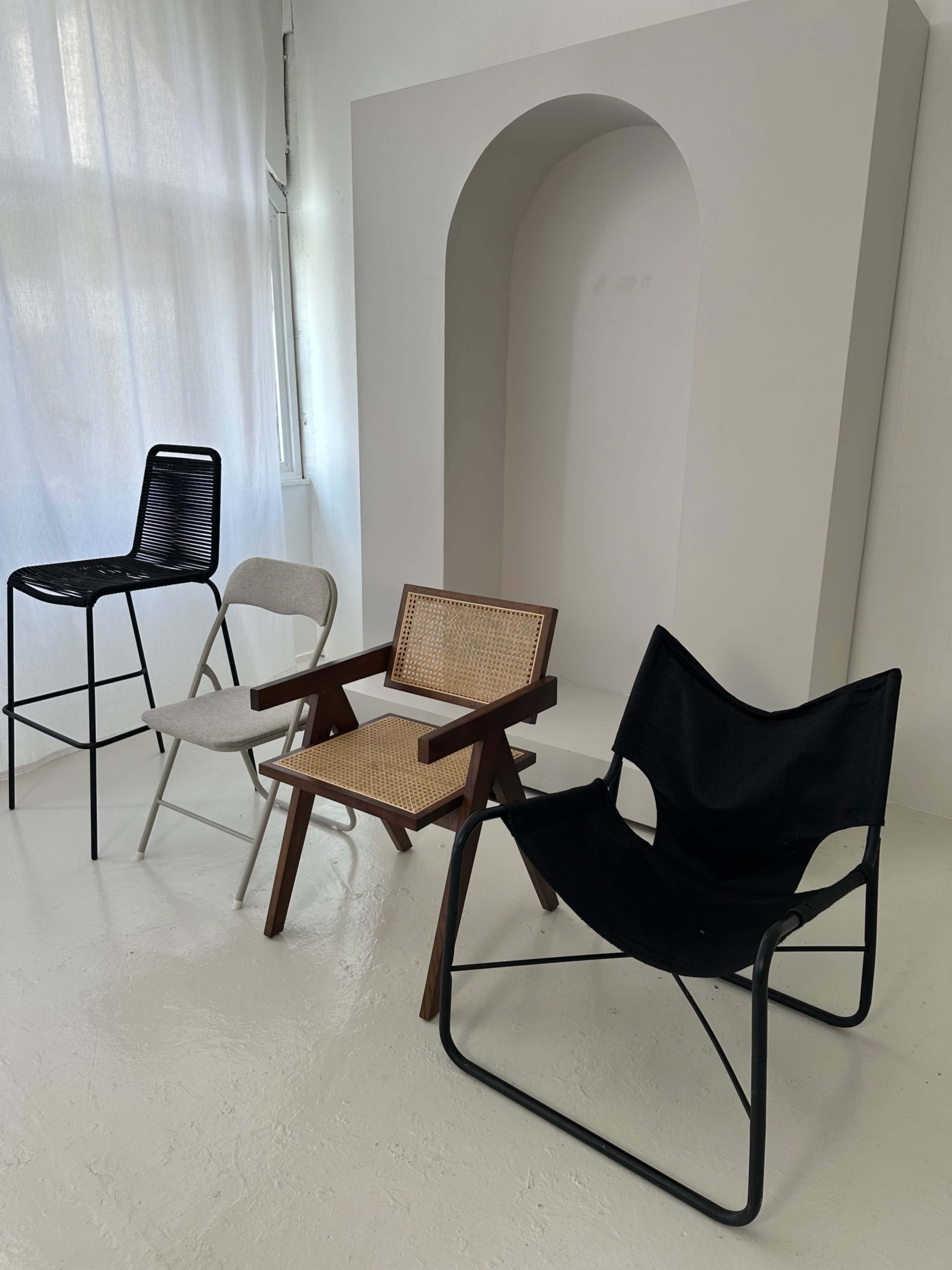 The image shows four different chairs arranged in a minimalist interior space with a white backdrop.