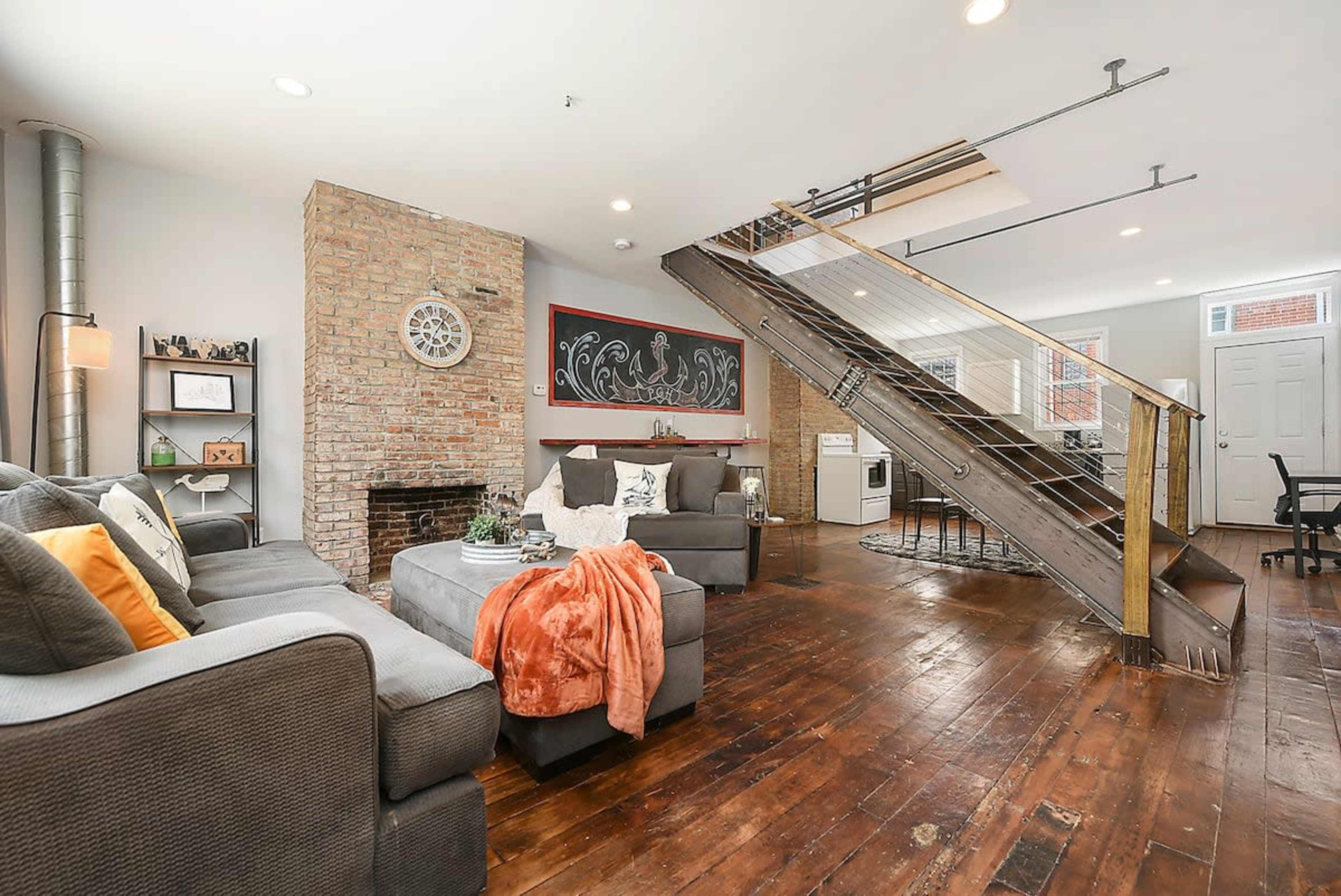 A modern living room with a brick wall, a gray couch, a fireplace, and a staircase leading to an upper level.