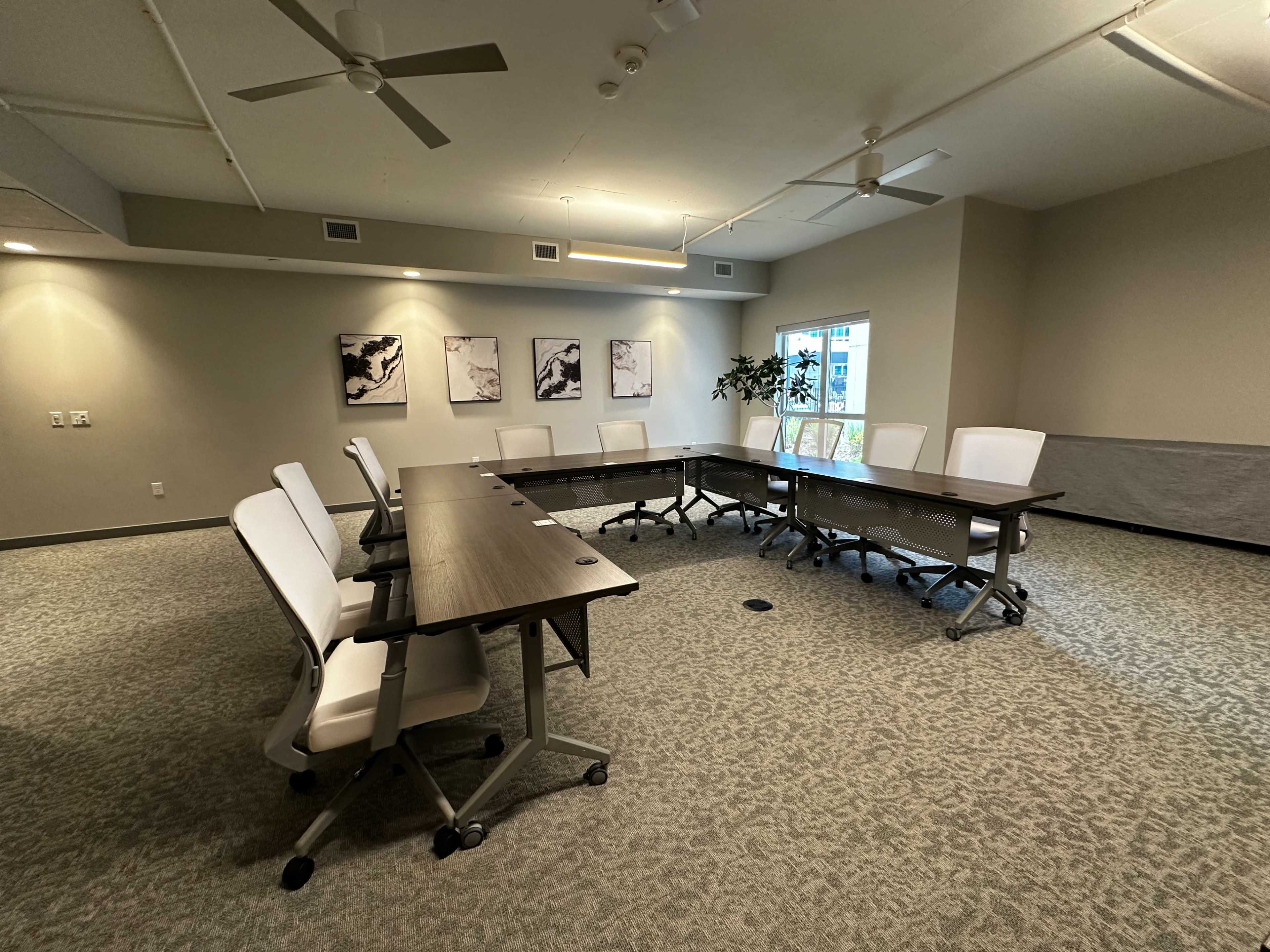 A spacious, well-lit conference room features a large rectangular table surrounded by white chairs, with framed artwork on the walls and a window that allows natural light to enter.