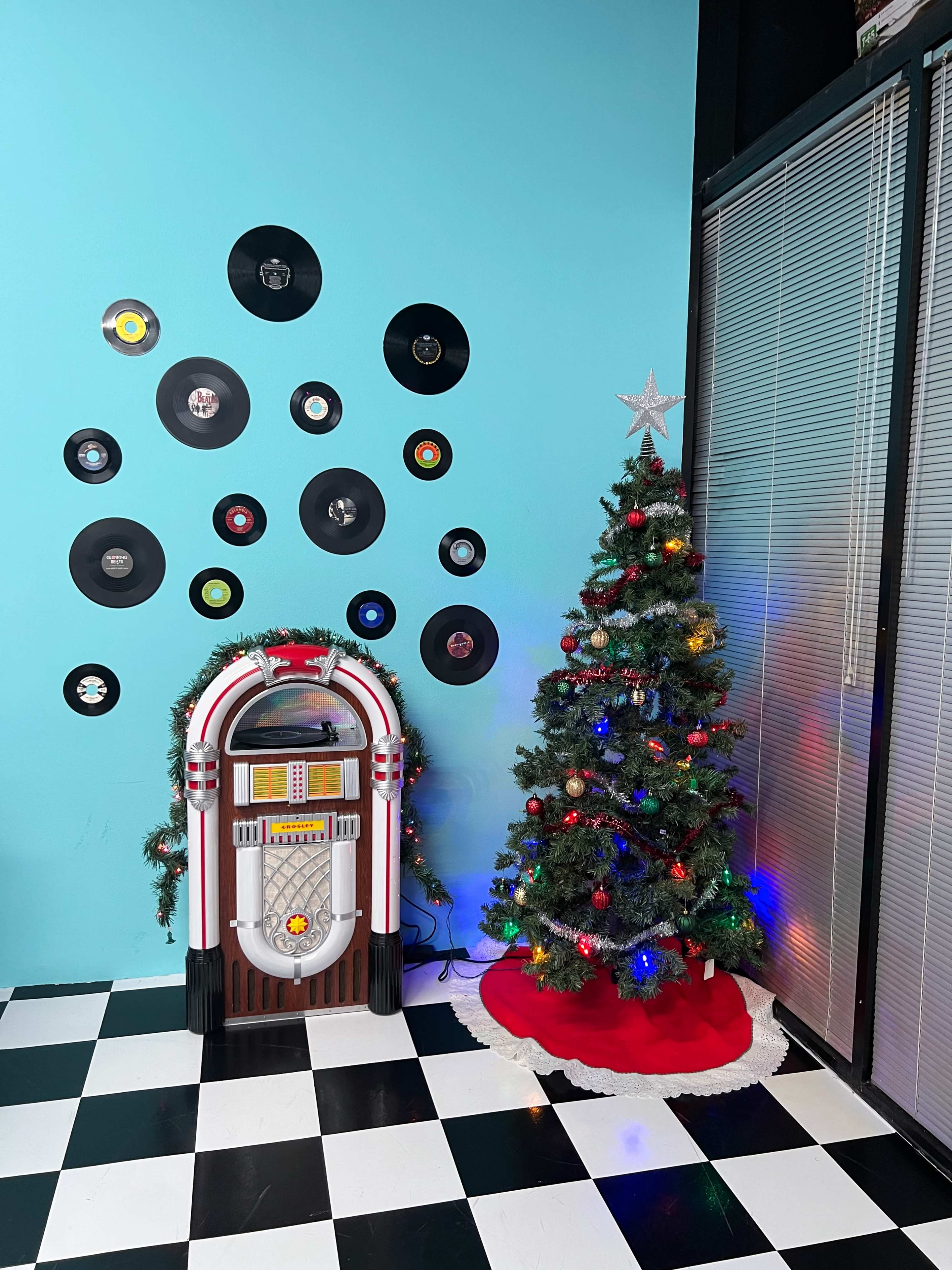 The image shows a retro jukebox beside a decorated Christmas tree against a bright blue wall, with vinyl records mounted decoratively above.