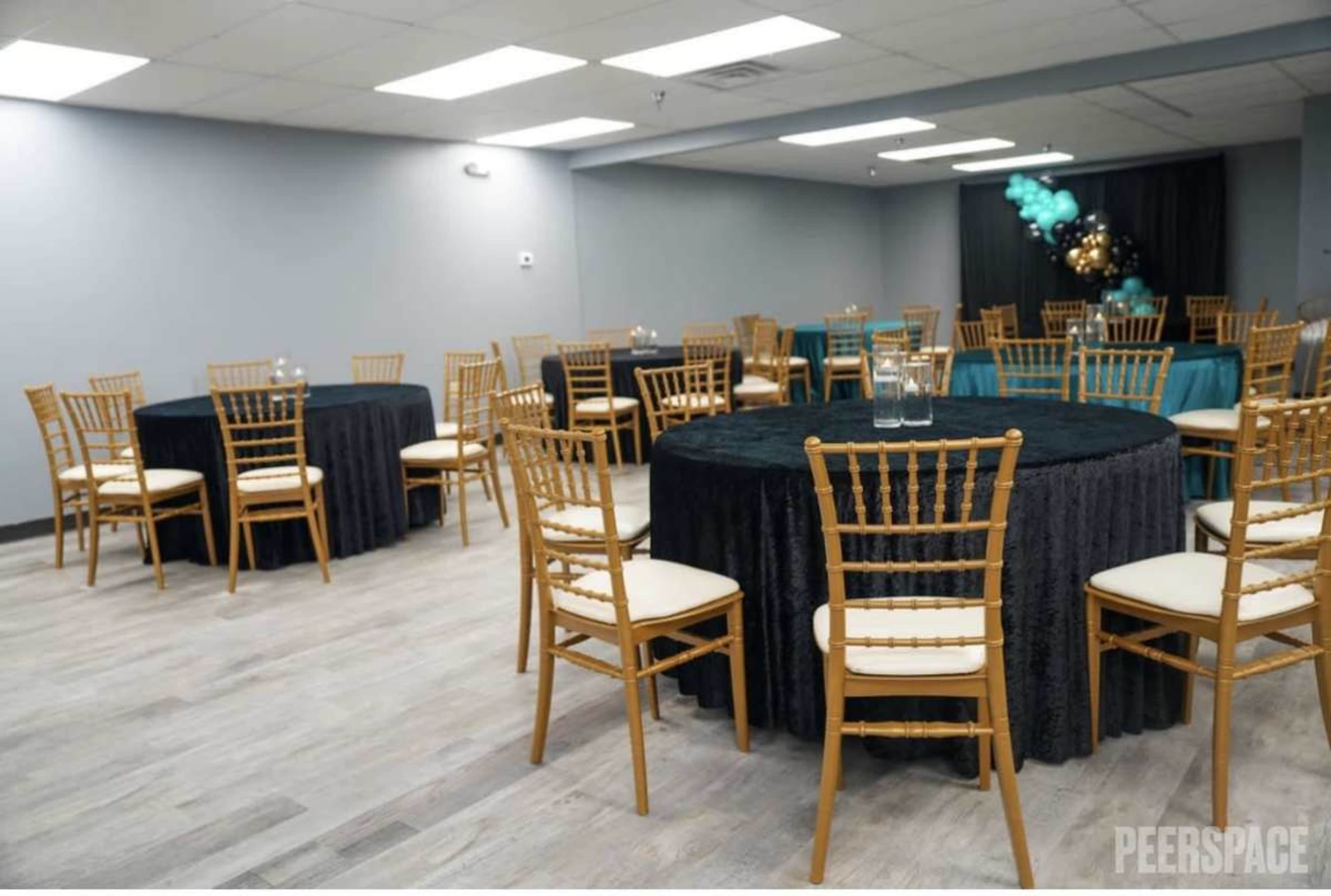 The image shows a spacious event room with several round tables covered in black and teal tablecloths, accompanied by wooden chairs arranged around them.