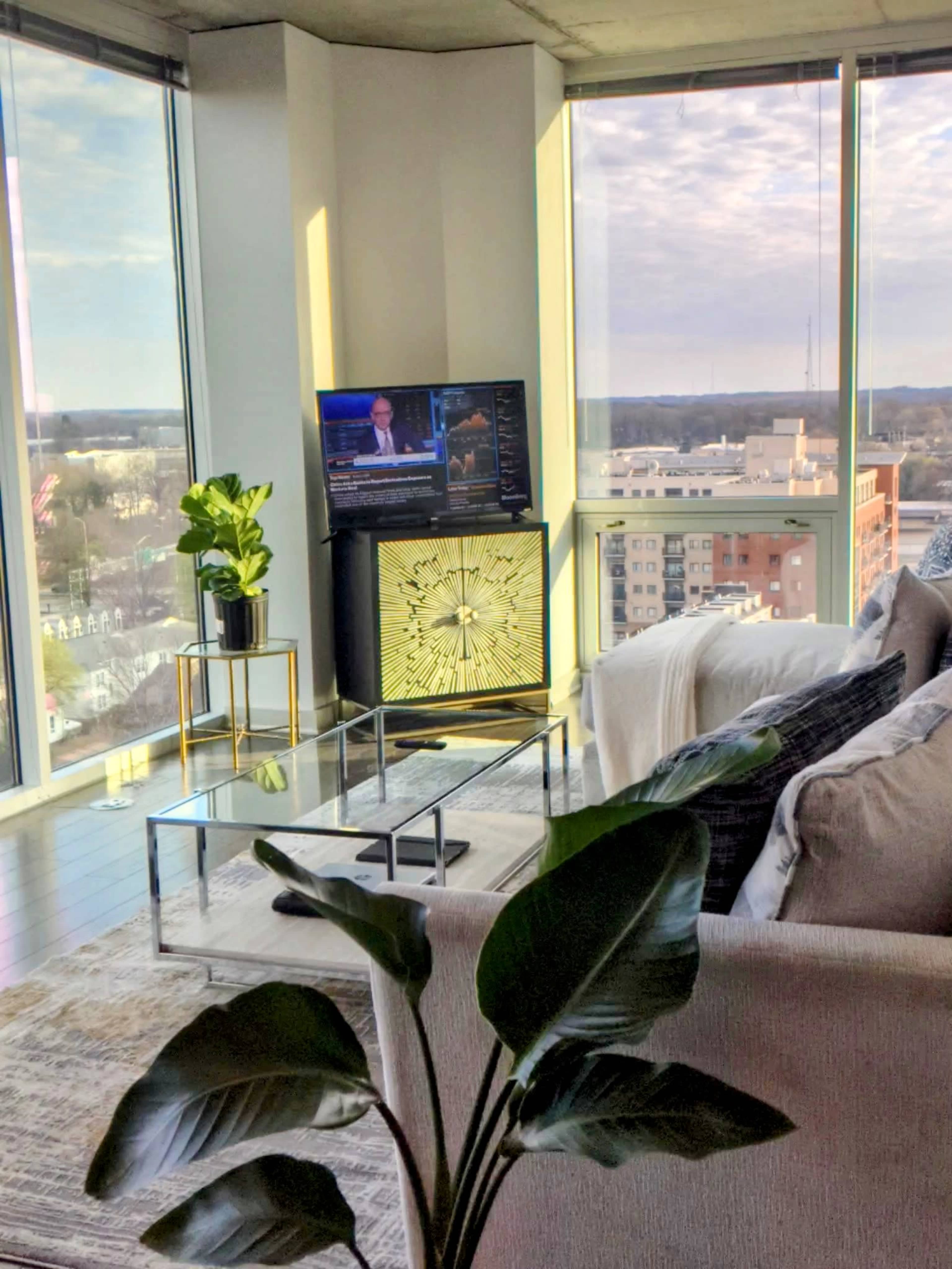 A modern living room features a glass coffee table, a light-colored couch, a decorative plant, and a television displaying news in front of large windows with city views.