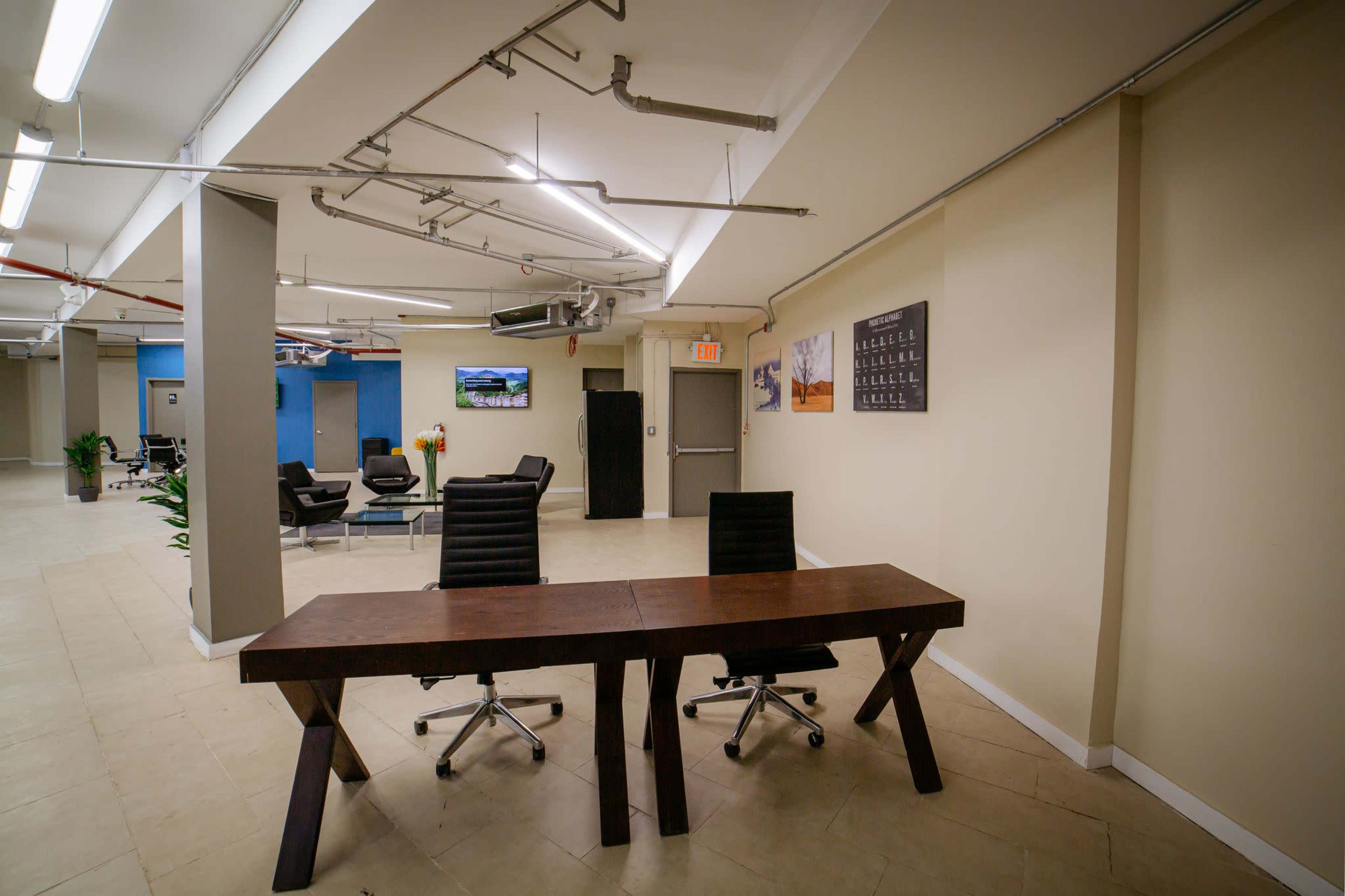 The image shows a modern office space featuring a wooden reception desk with two black chairs, surrounded by seating areas and exposed ductwork on the ceiling.