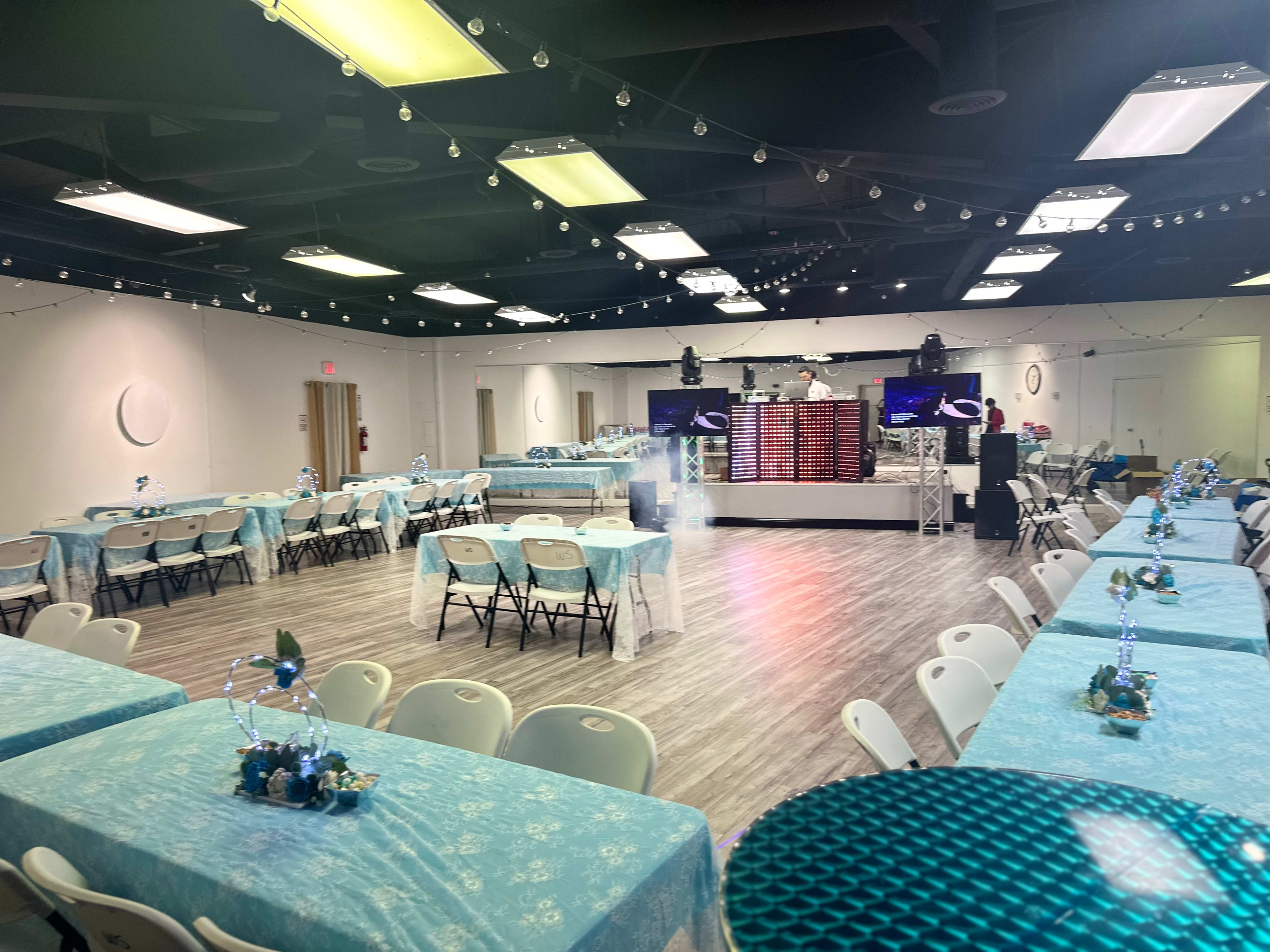 The image shows a spacious event hall set up for a celebration, featuring multiple tables with light blue tablecloths and a stage area with sound equipment and screens.
