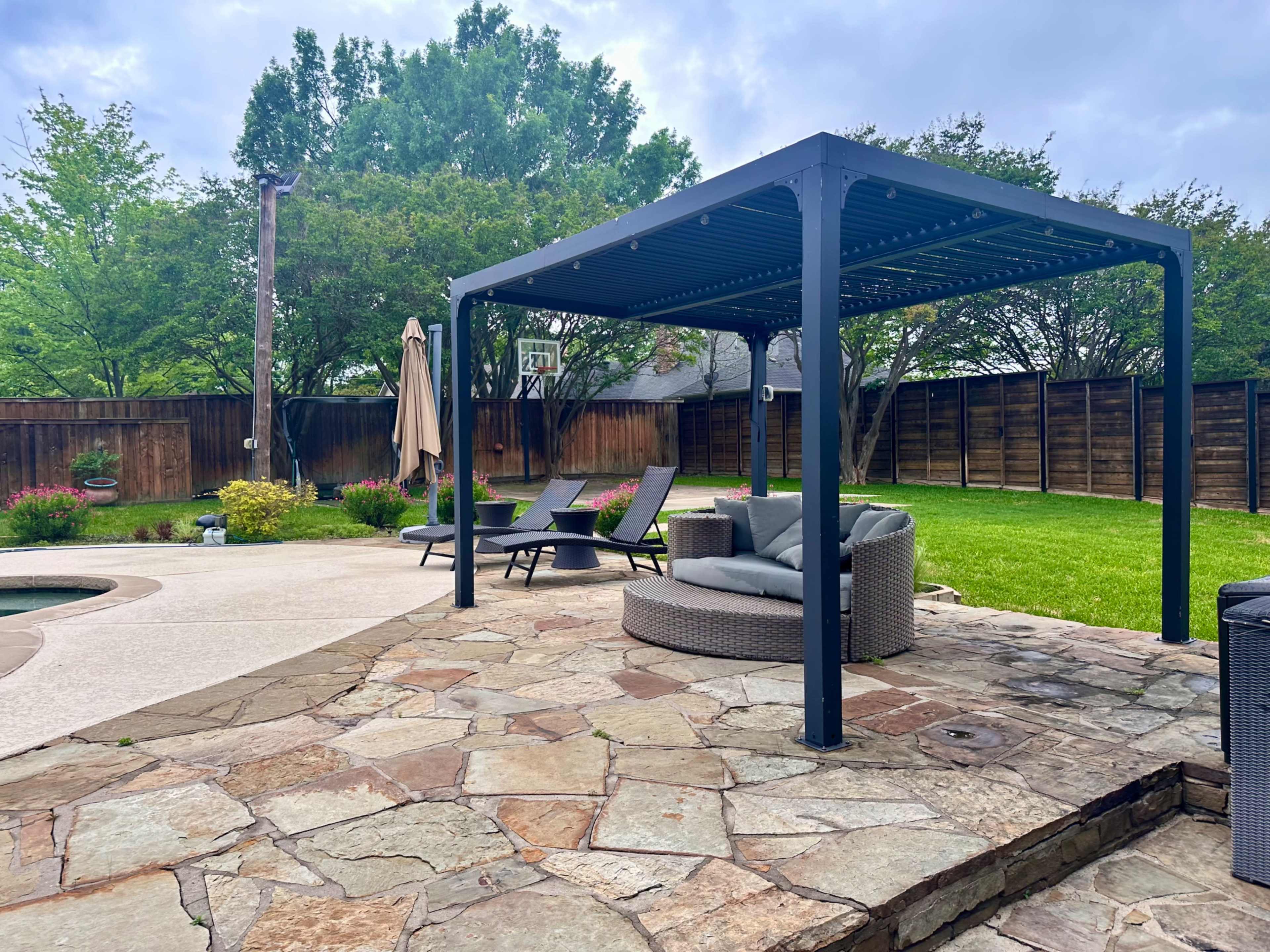 A stone patio features a shaded pergola with seating arrangements, surrounded by green grass and trees.