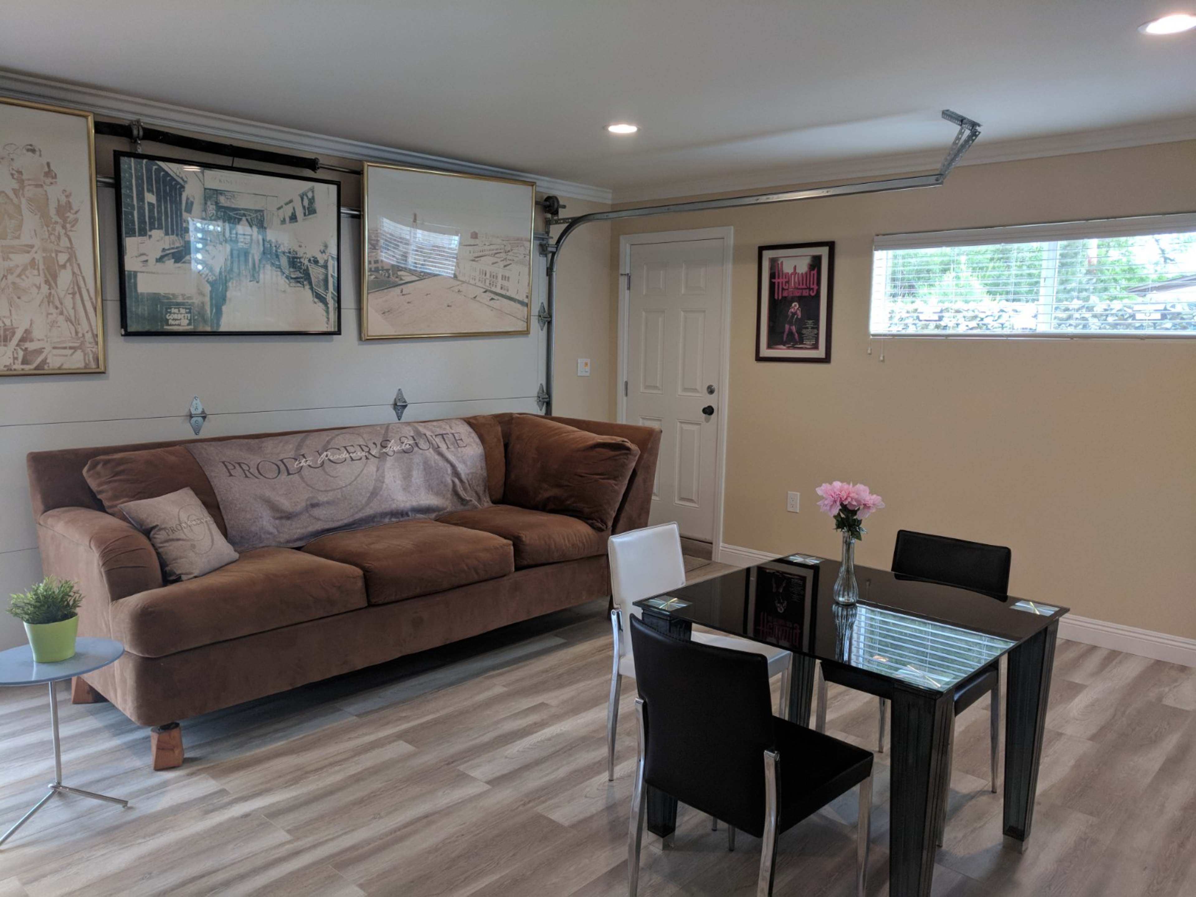 The image shows a cozy living space featuring a brown sofa, a glass dining table with black chairs, and wall art in a well-lit room.
