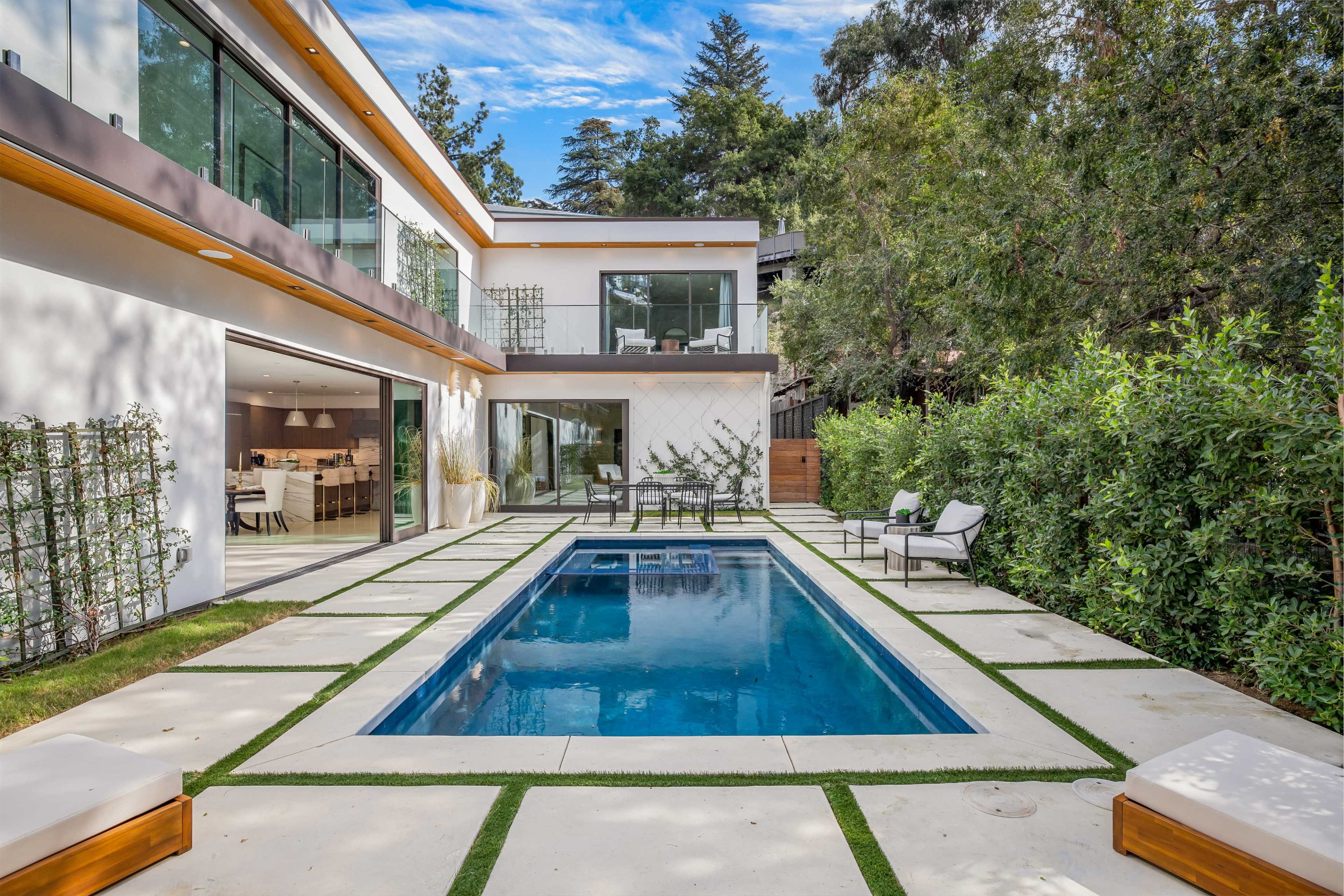 The image shows a modern house with a backyard featuring a rectangular swimming pool surrounded by concrete tiles and green landscaping.