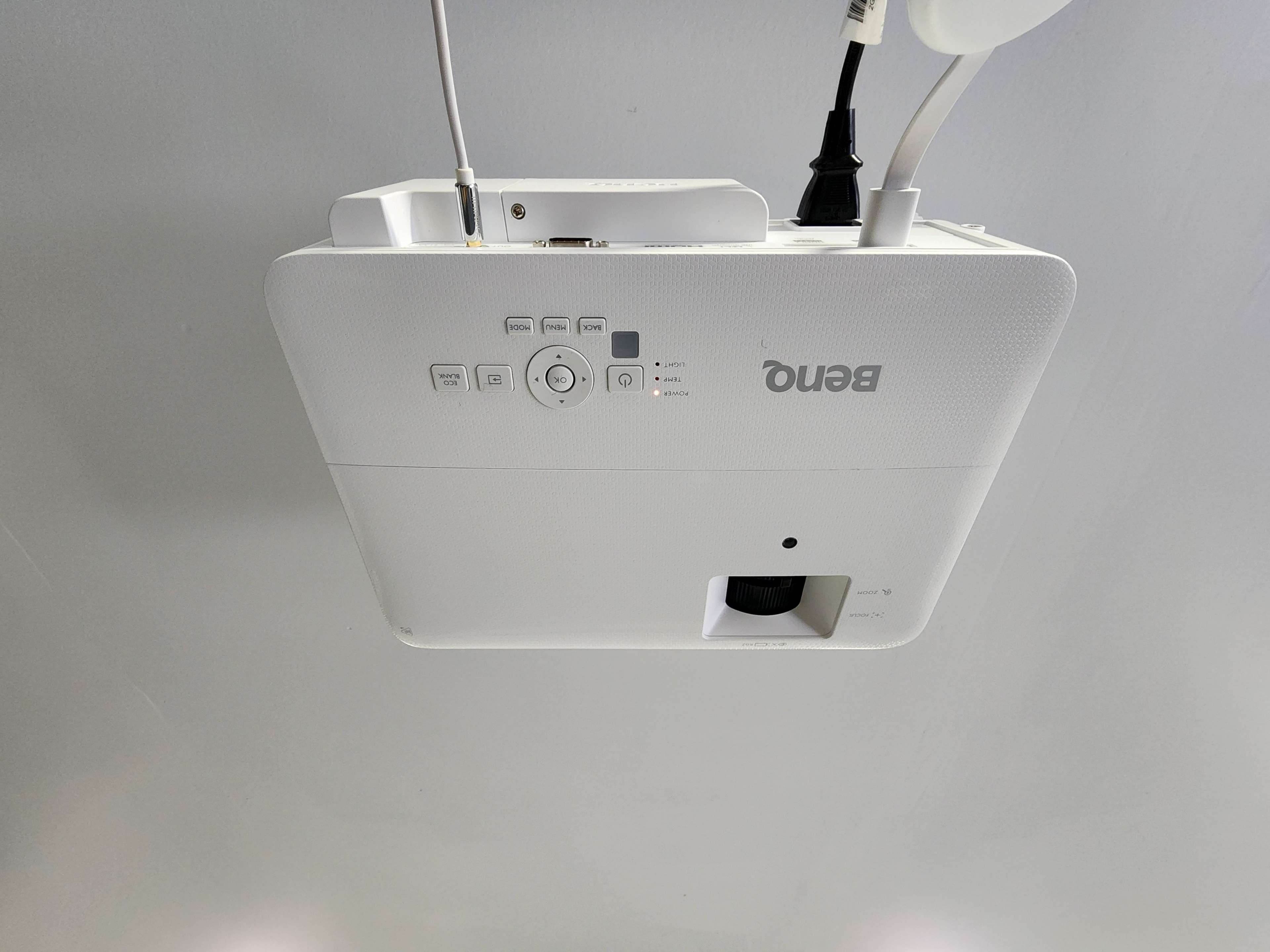 A white BenQ projector mounted on a ceiling, with various control buttons and a cable connected to it.