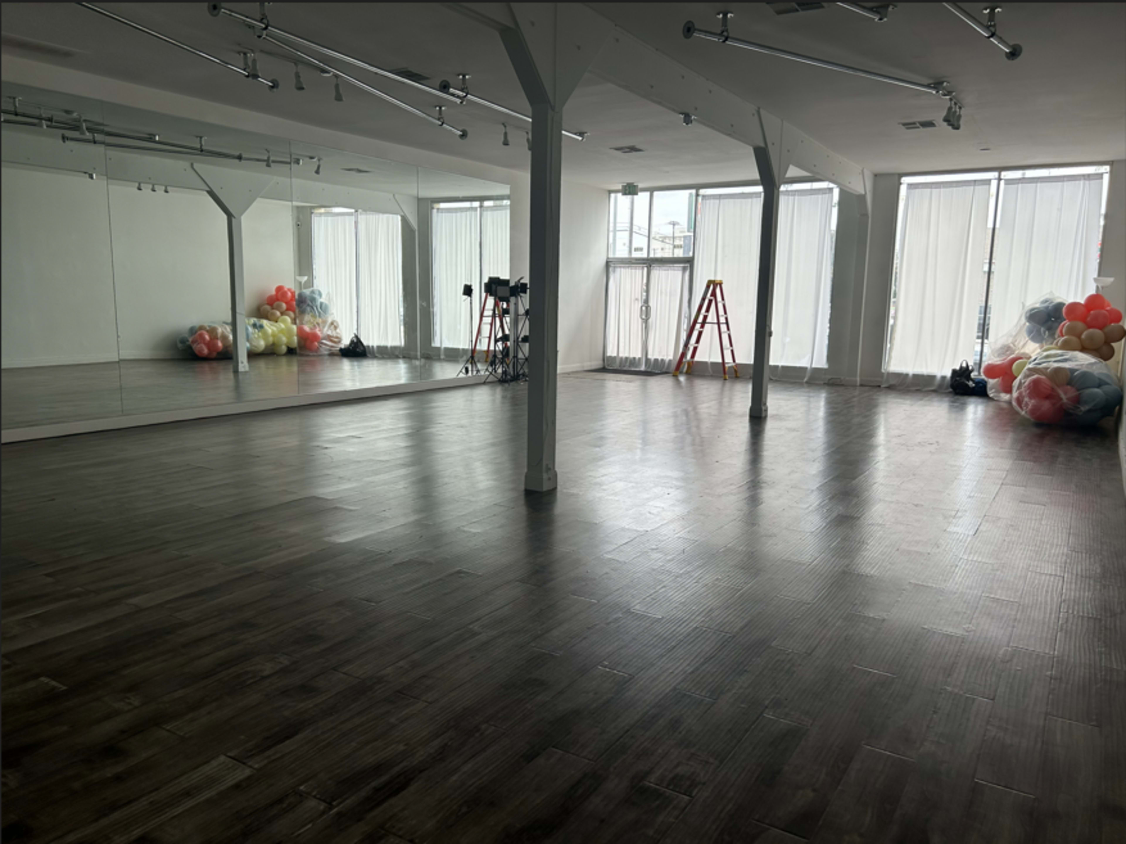 The image shows a spacious, well-lit studio with large windows, a mirrored wall, and a collection of colorful balloons piled in one corner.
