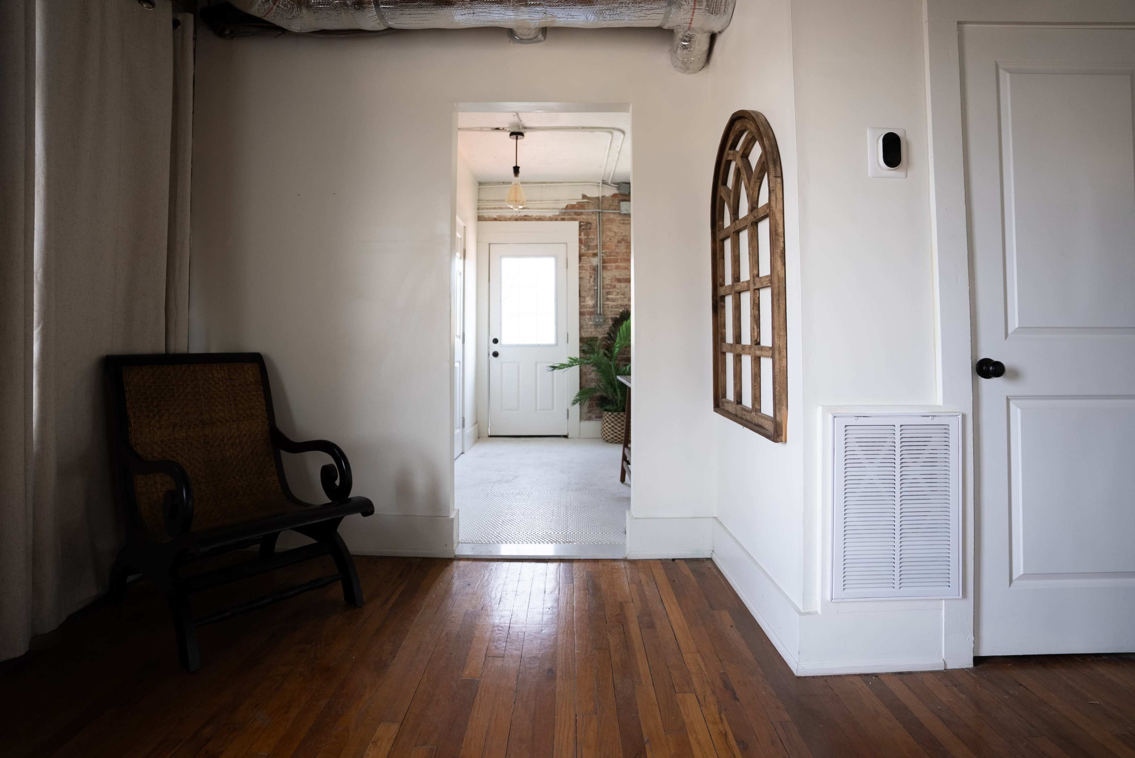 The image shows a hallway with a wooden chair, a decorative window frame on the wall, and a door leading to a room with plants visible through the open doorway.