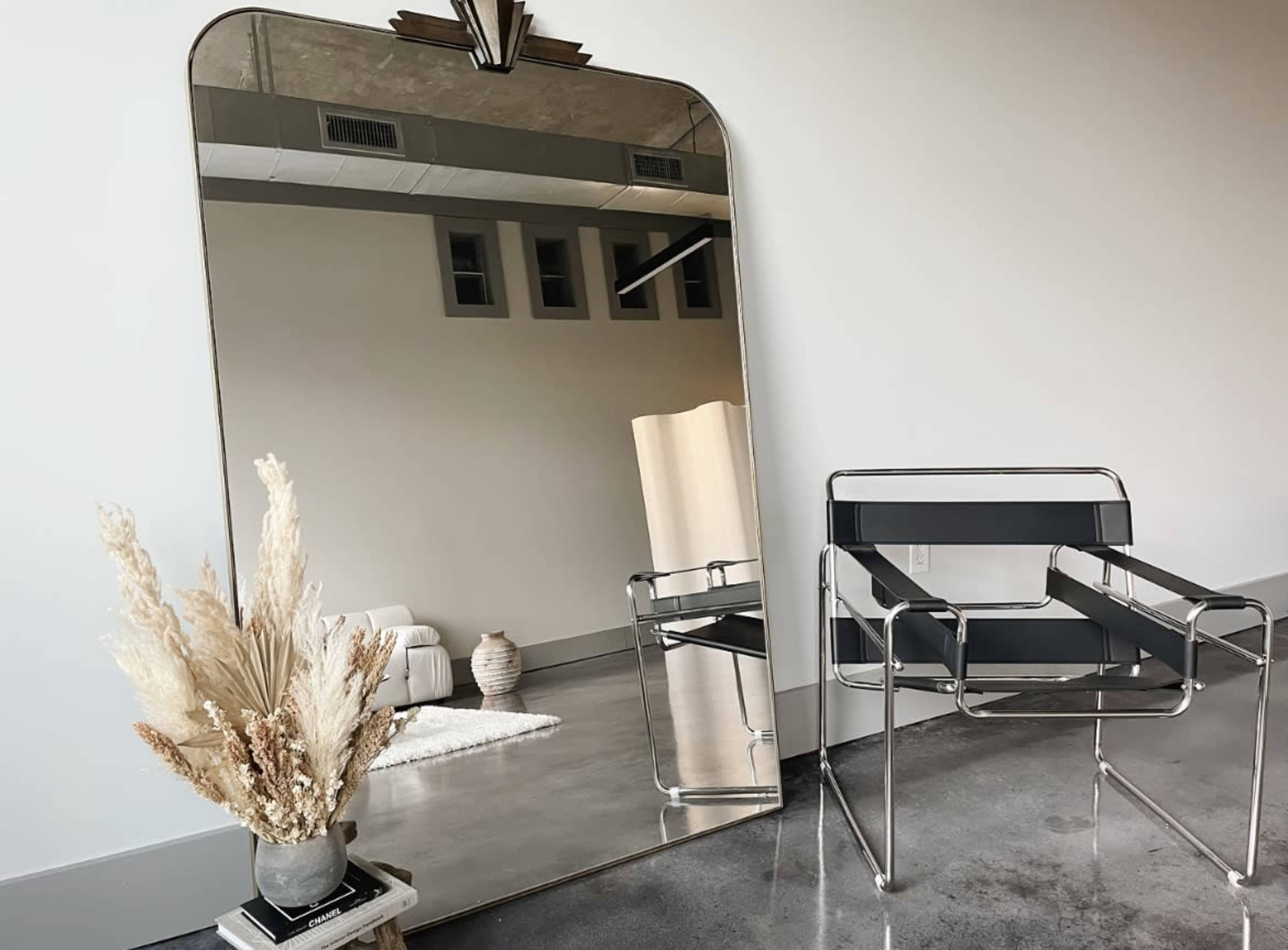 A large mirror with a decorative top stands next to a sleek black chair and a vase of dried flowers in a minimalistic room.