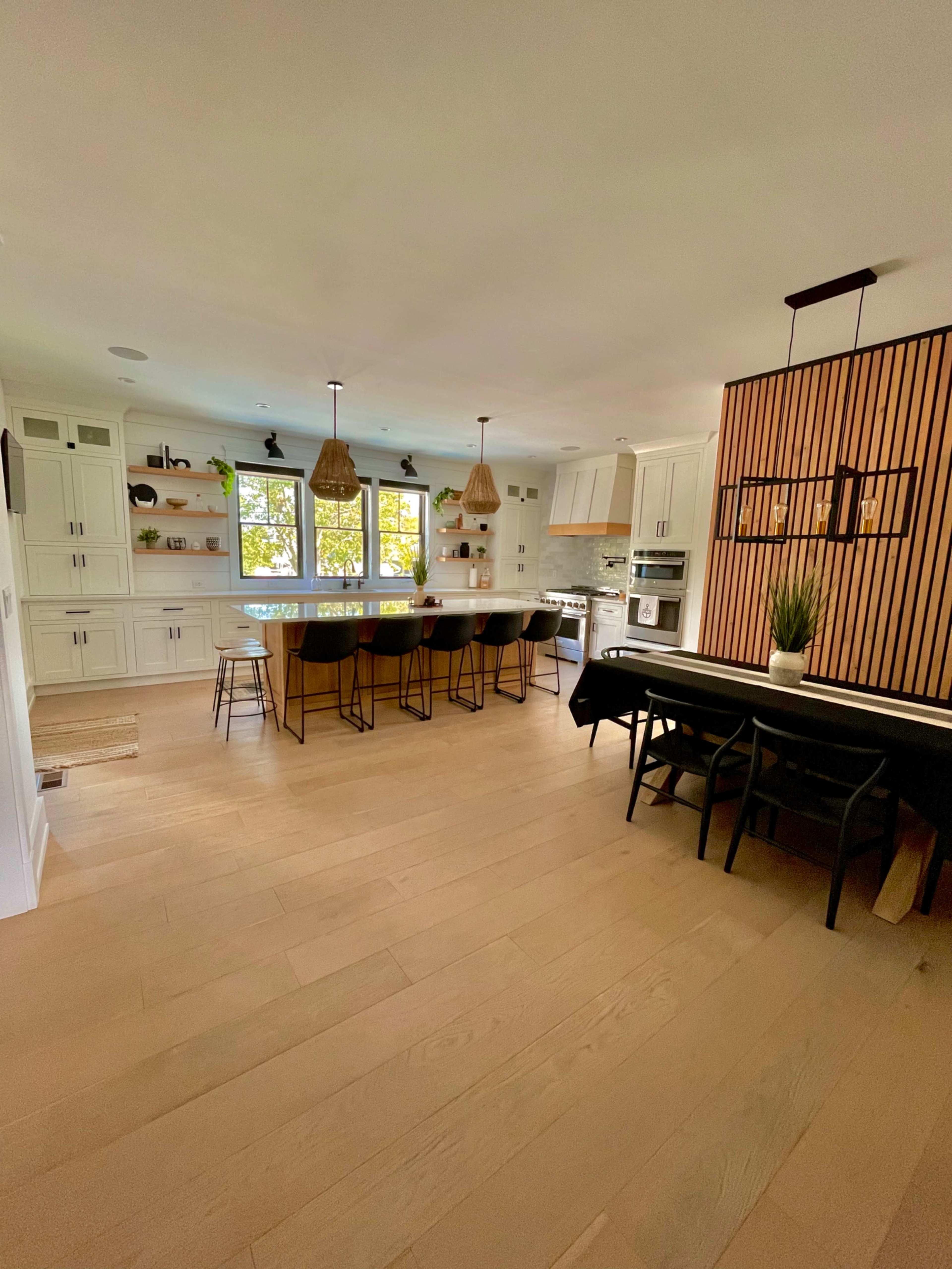 A modern kitchen with white cabinetry, a large island with bar stools, and a dining table along one side, illuminated by pendant lights.