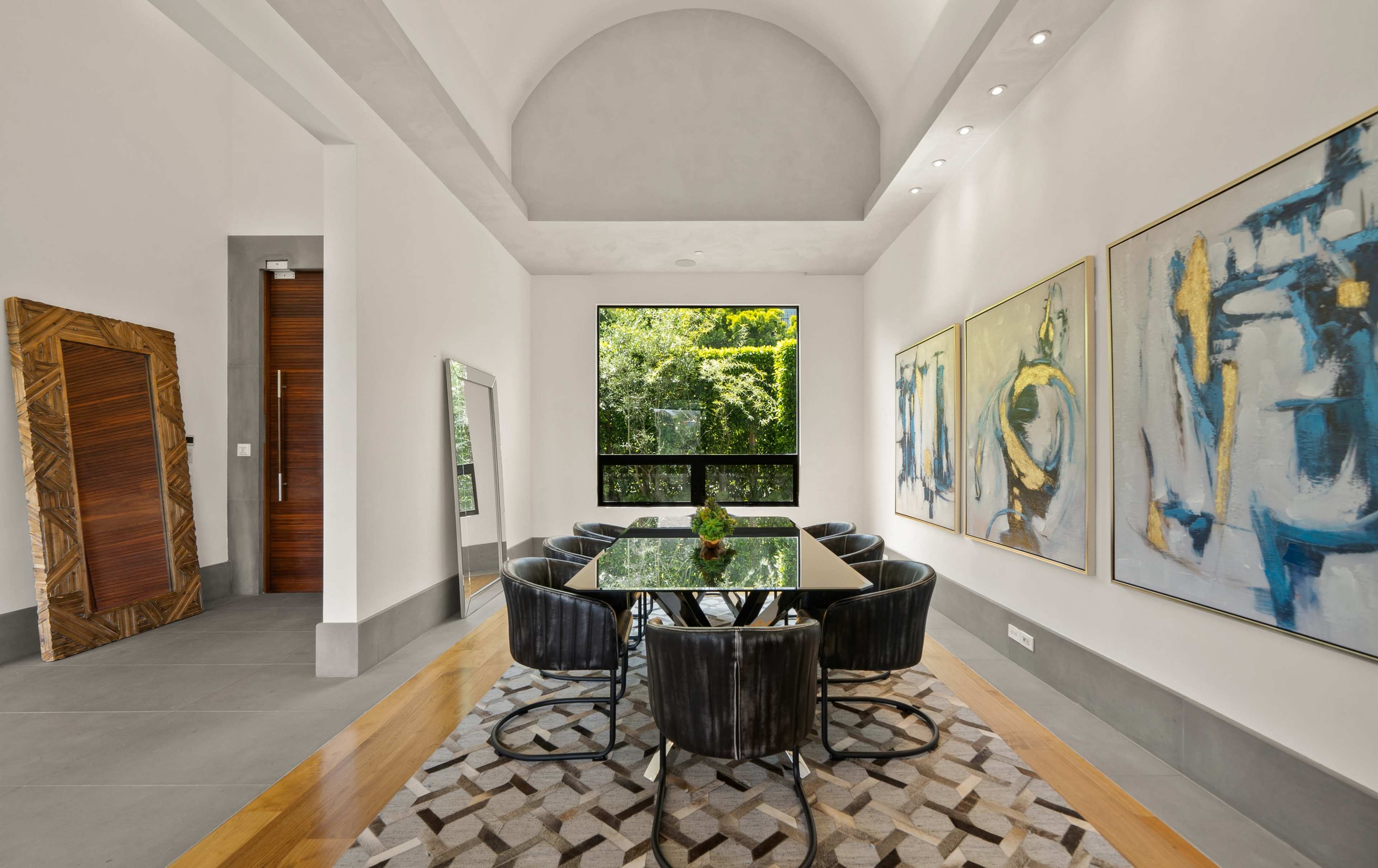 A modern dining room features a glass-top table surrounded by black chairs, with large abstract paintings on the walls and a view of greenery through a large window.