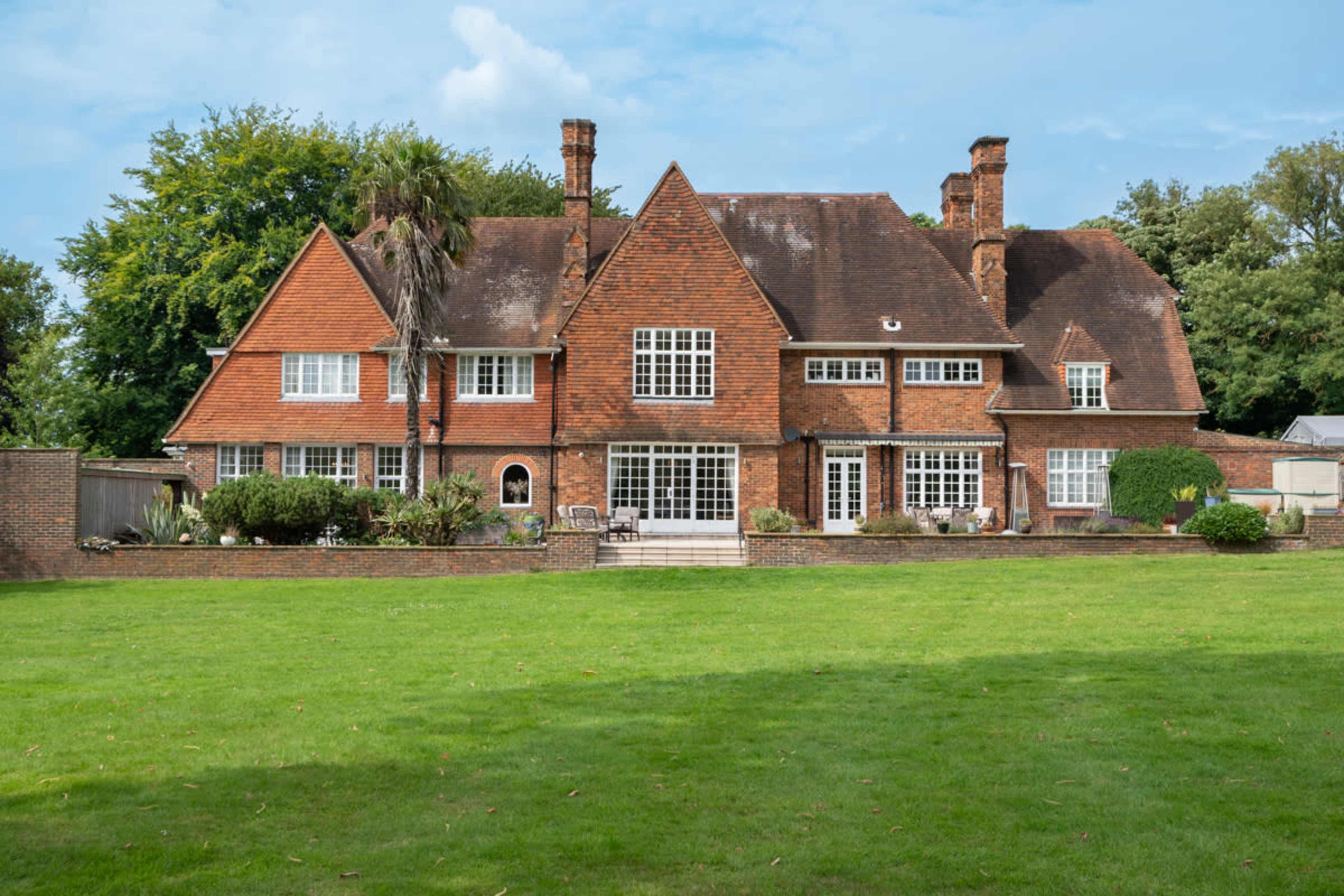 A large brick house with multiple gabled roofs and a well-maintained lawn is set against a backdrop of trees.