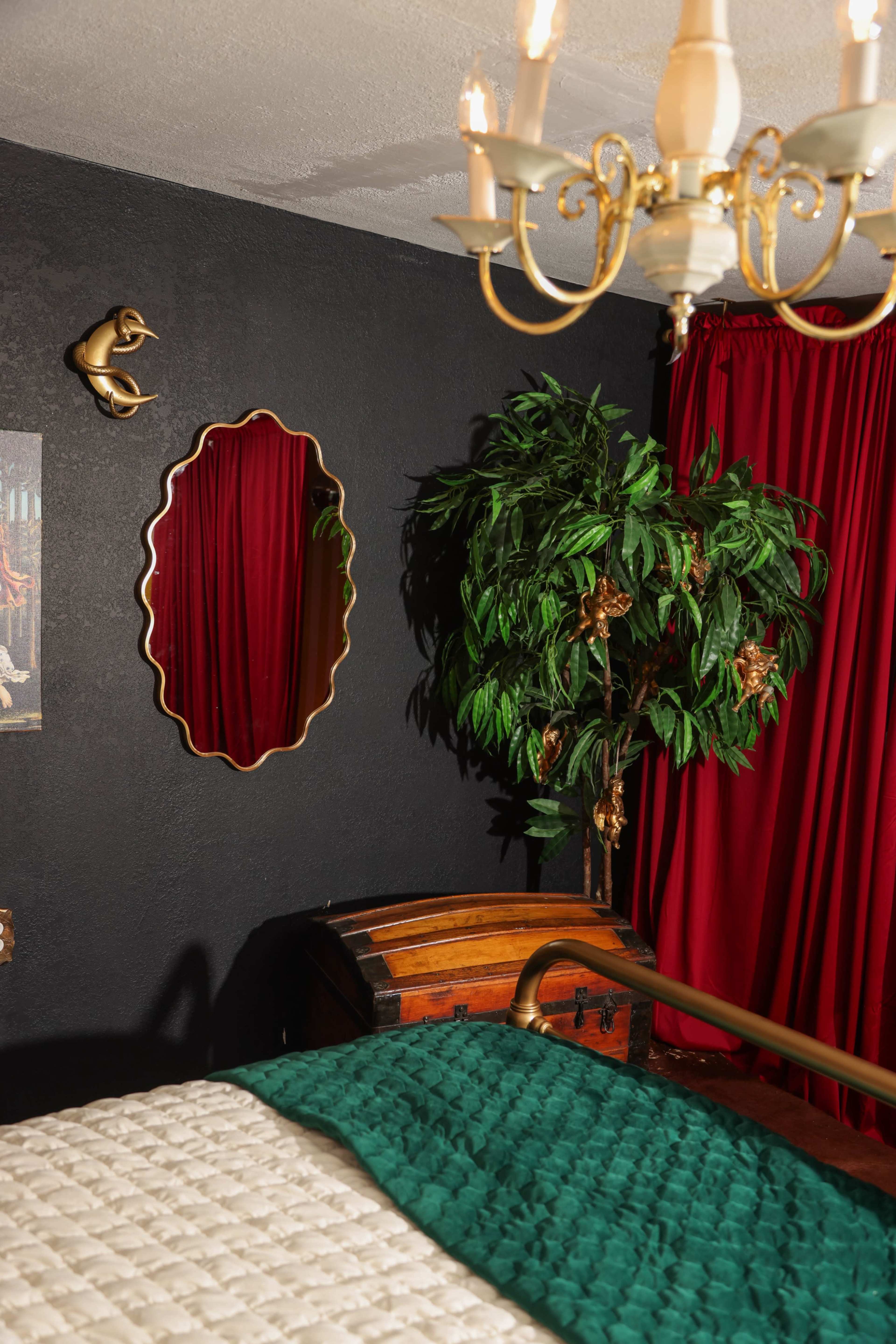 The image shows a dark bedroom with a brass chandelier, a large red curtain, a decorative mirror, a potted plant, and a wooden trunk at the foot of the bed.