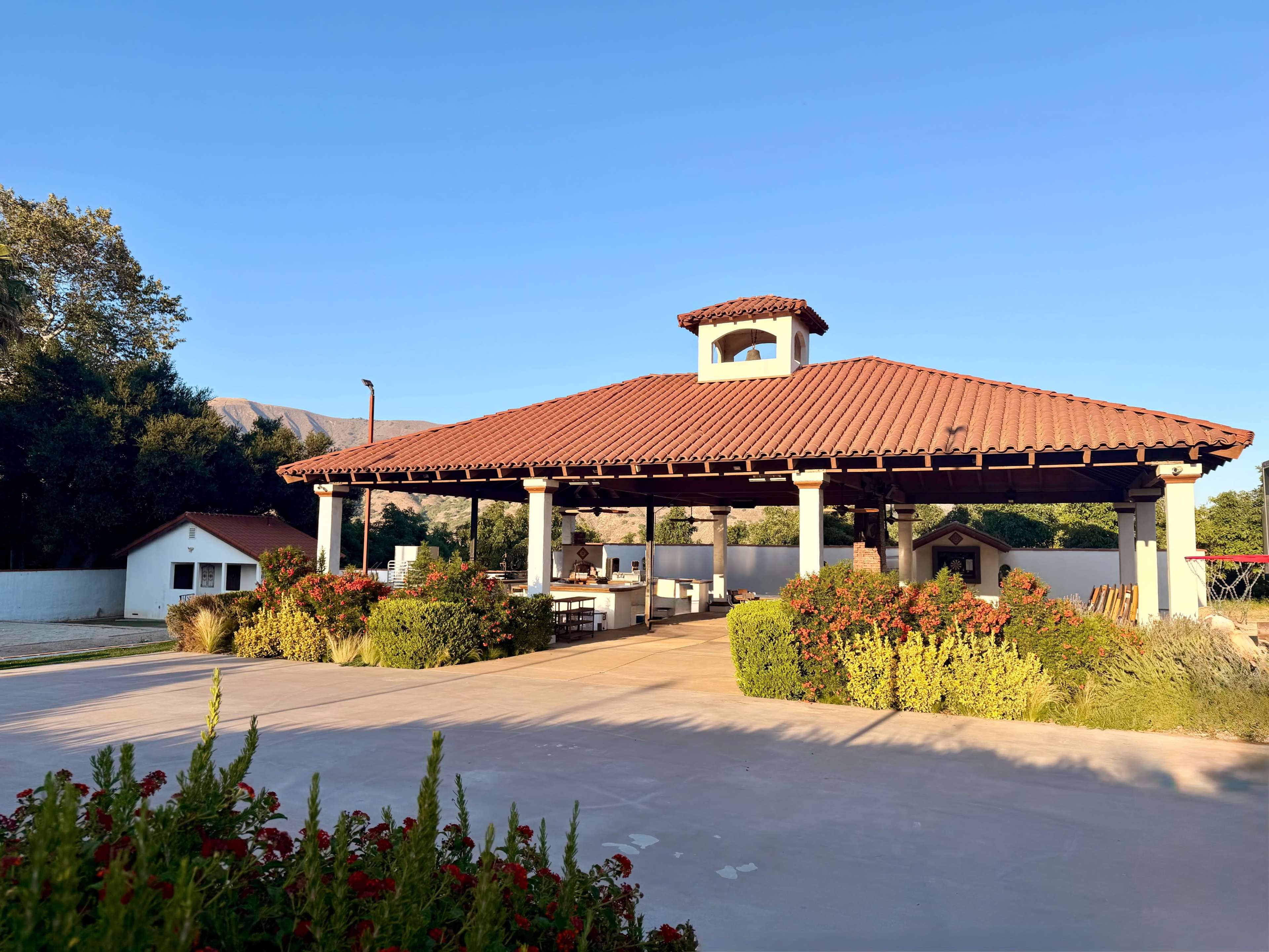 The image shows a large open pavilion with a tiled roof, set against a clear blue sky, surrounded by landscaped gardens and a small building in the background.