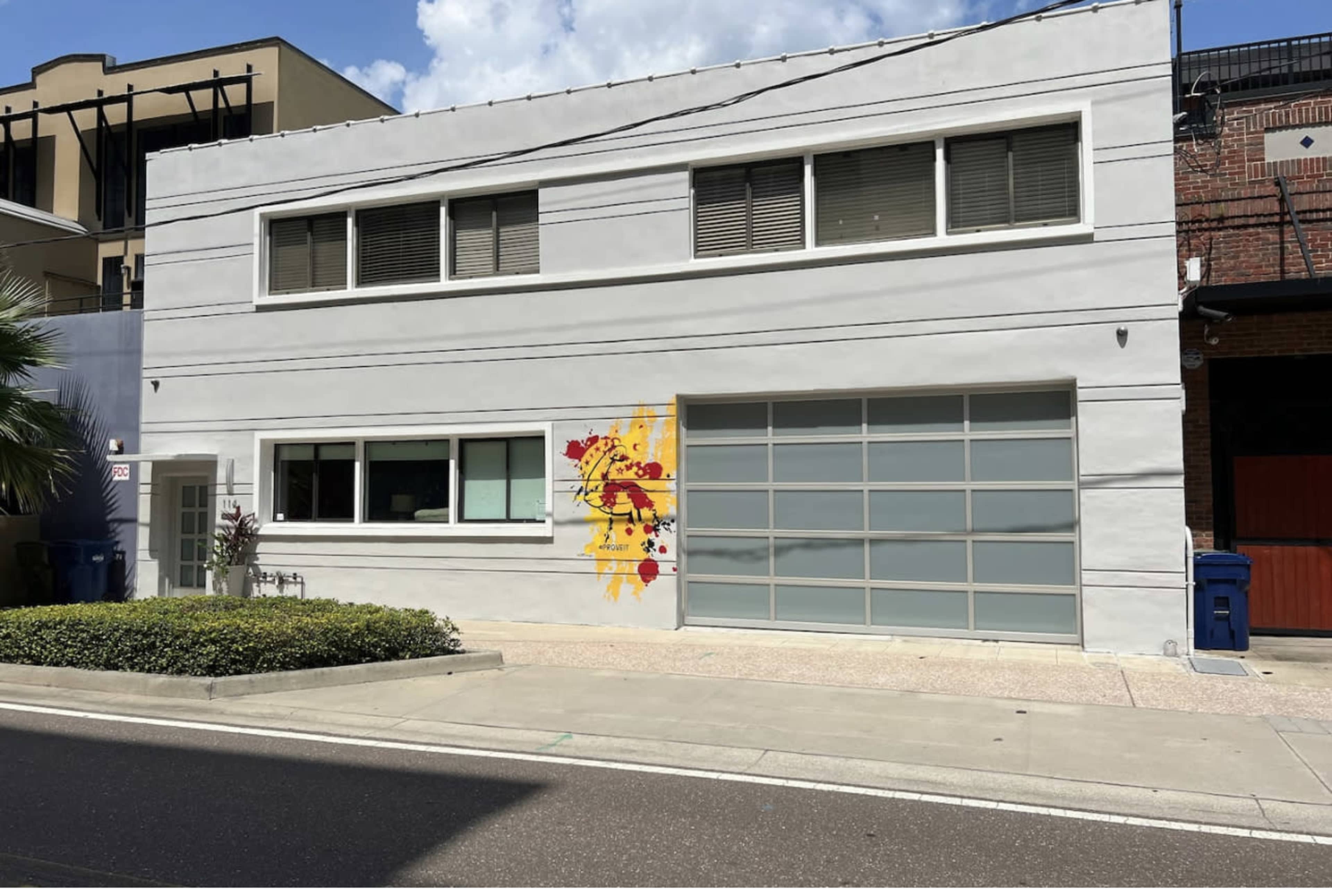 A modern gray building with large windows and a colorful mural on one side, located on a clean street with parked bins.