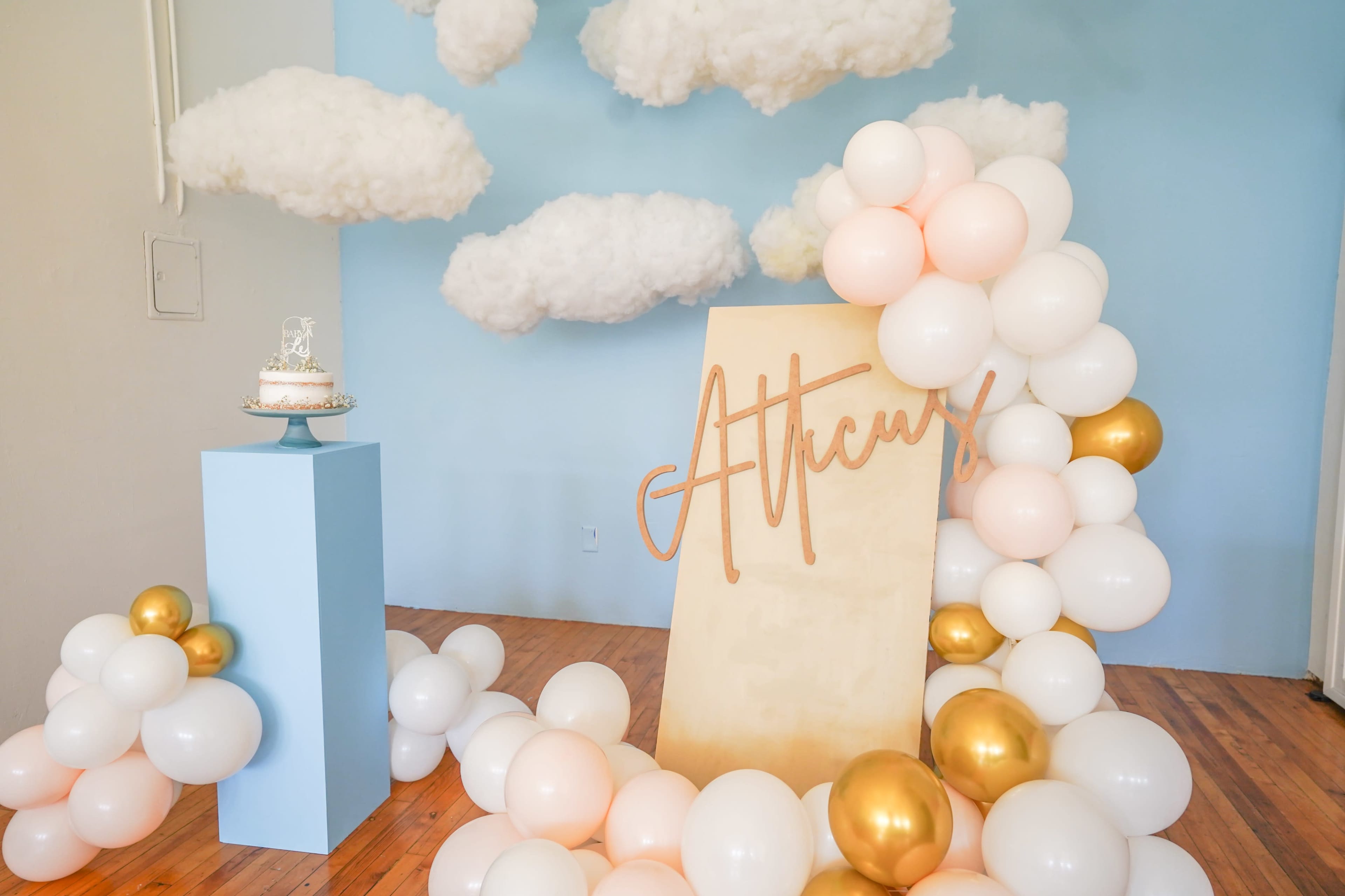 A decorative display featuring a cake on a blue pedestal, surrounded by an arrangement of balloons in pink, white, and gold, with a wooden sign that reads "Atticus" and fluffy cloud decorations against a light blue wall.