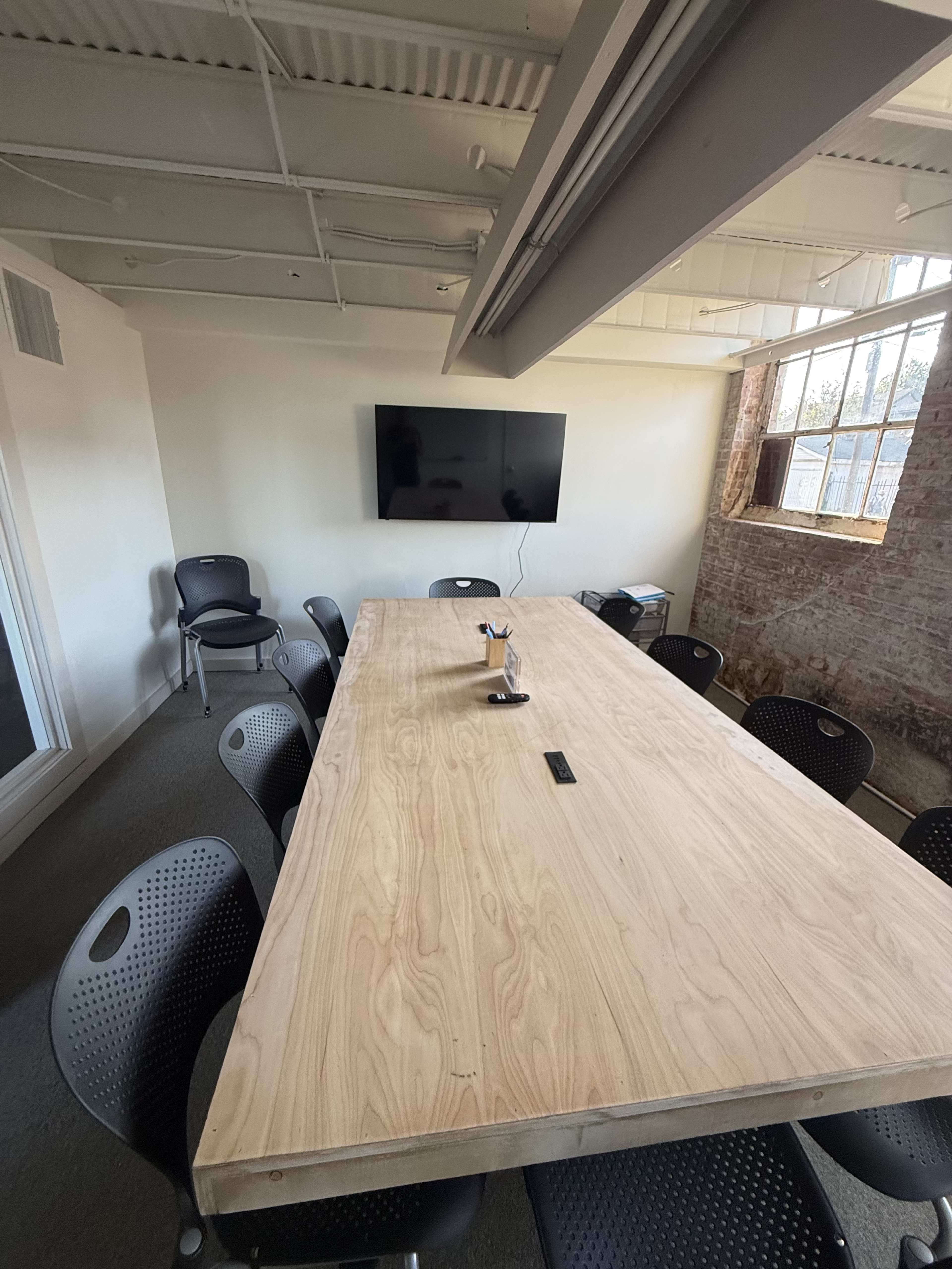 A long wooden conference table is surrounded by black chairs in a well-lit meeting room with a television mounted on the wall.