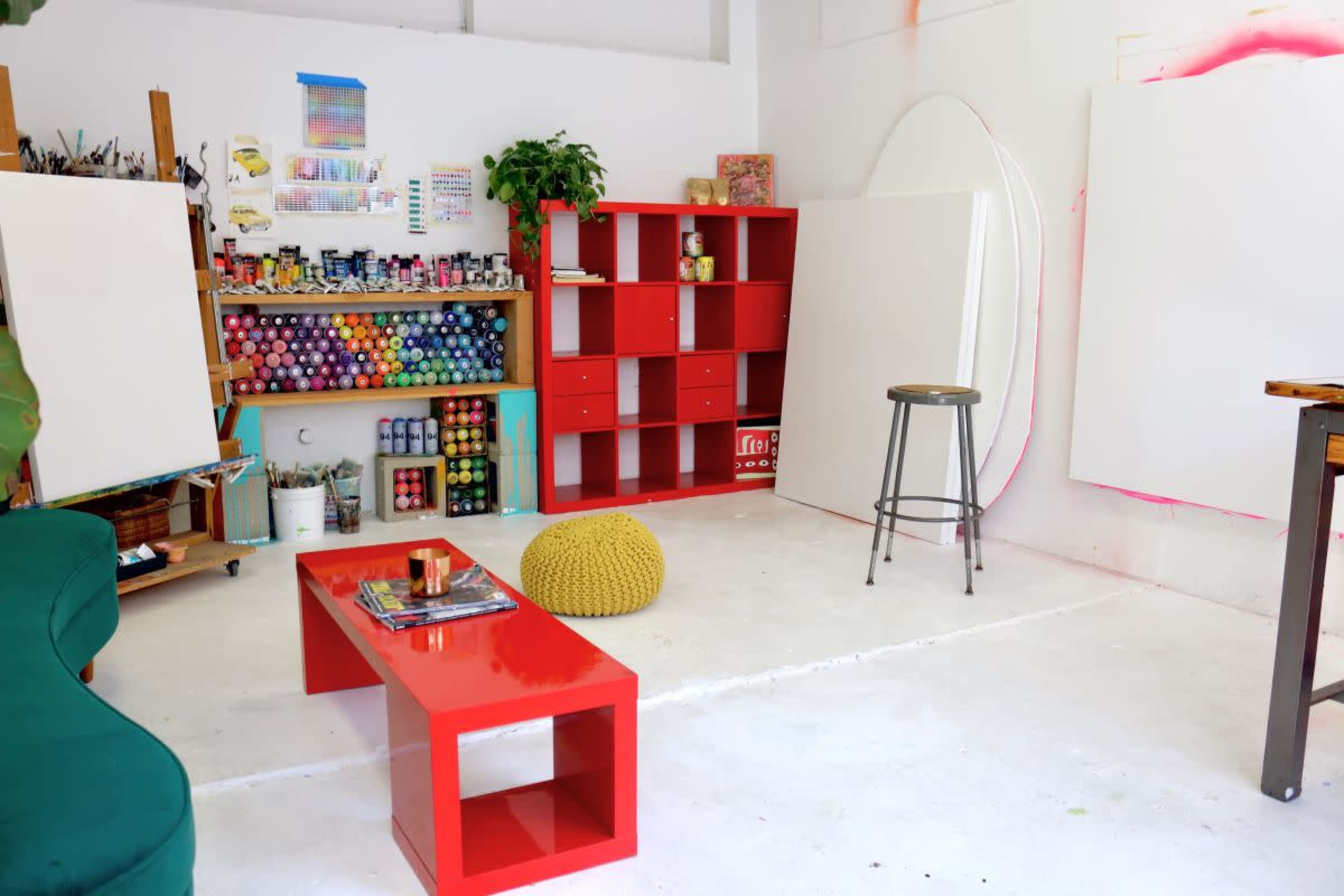 An art studio with a bright red shelving unit, organized paint supplies, a green couch, and empty canvases set against a white wall.