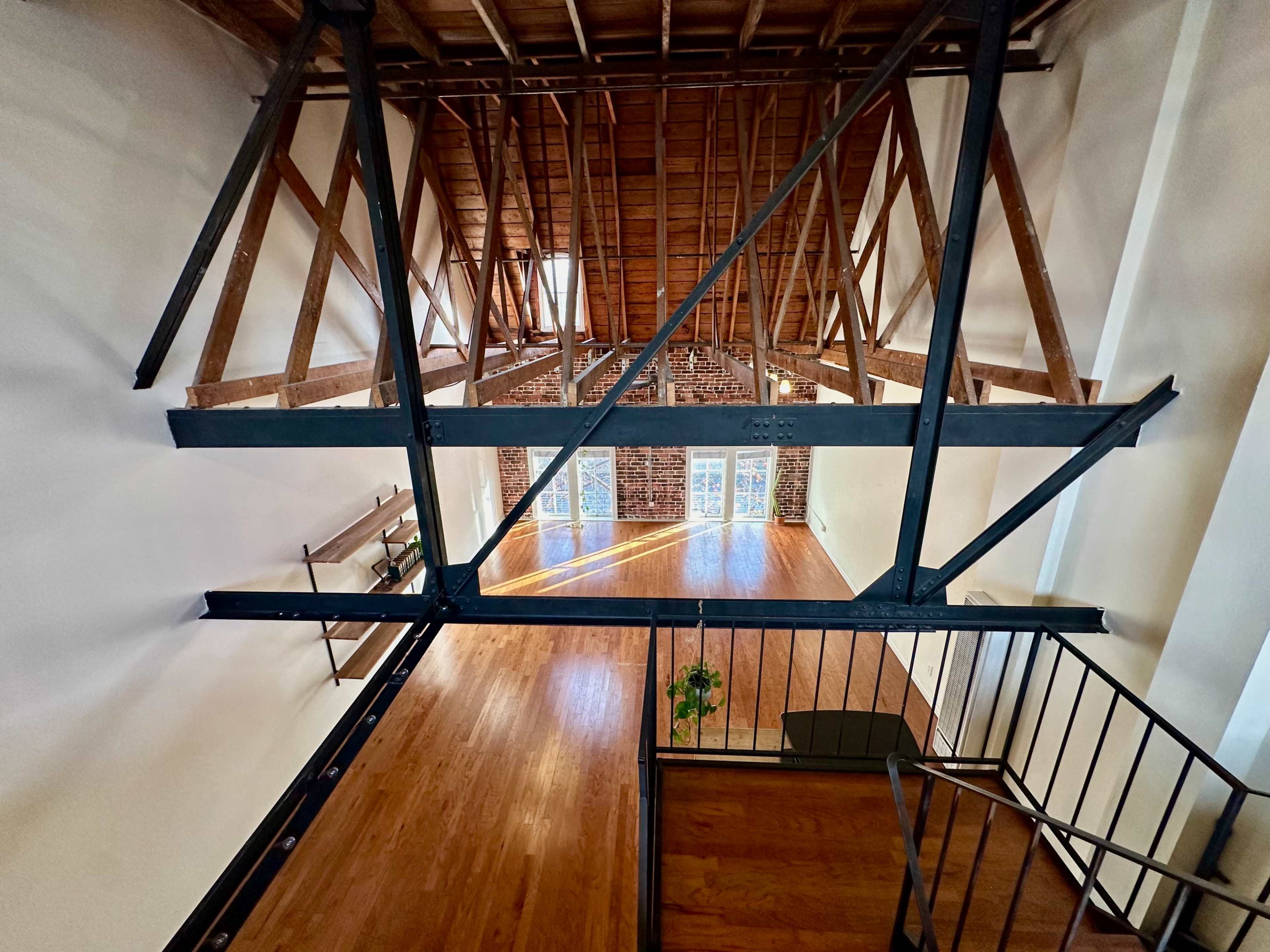The image shows an open loft space with wooden flooring, exposed brick walls, and a high ceiling supported by metal beams.