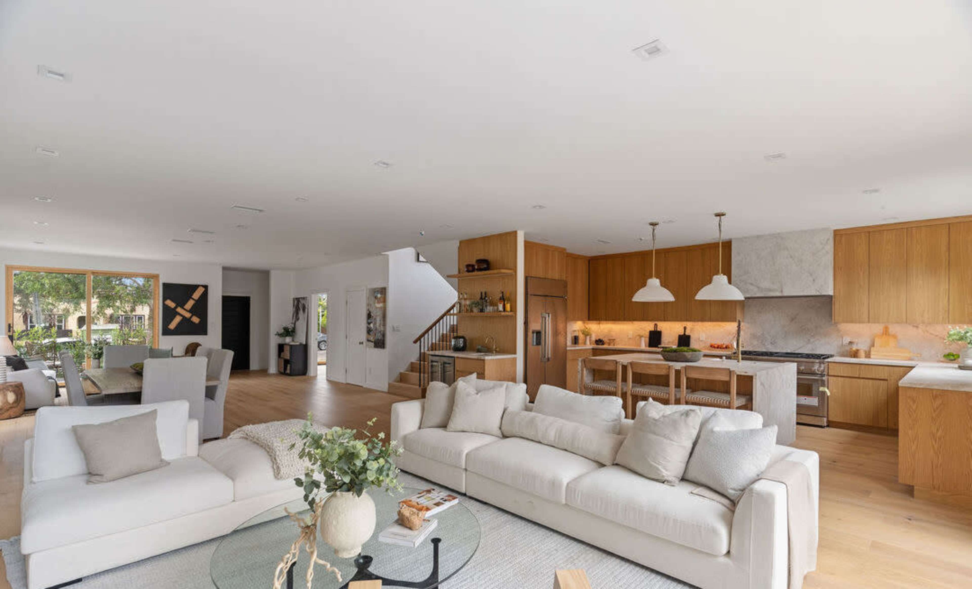 The image shows a modern, open-concept living space with a light-colored sofa, a round glass coffee table, and a kitchen featuring wooden cabinets and pendant lights.