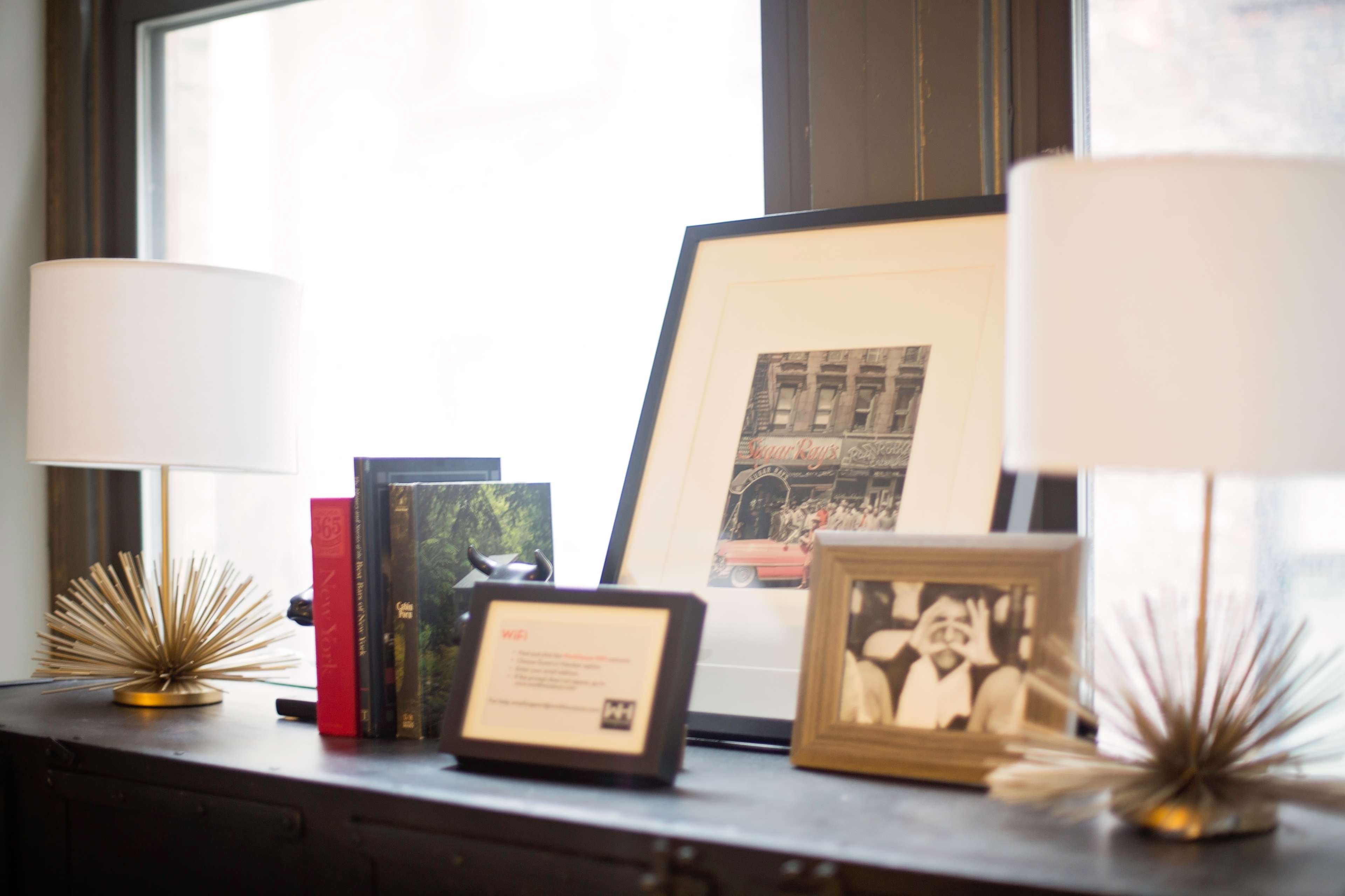 A tabletop with two table lamps, a framed photograph, a picture frame, several books, and an art print arranged near a window.