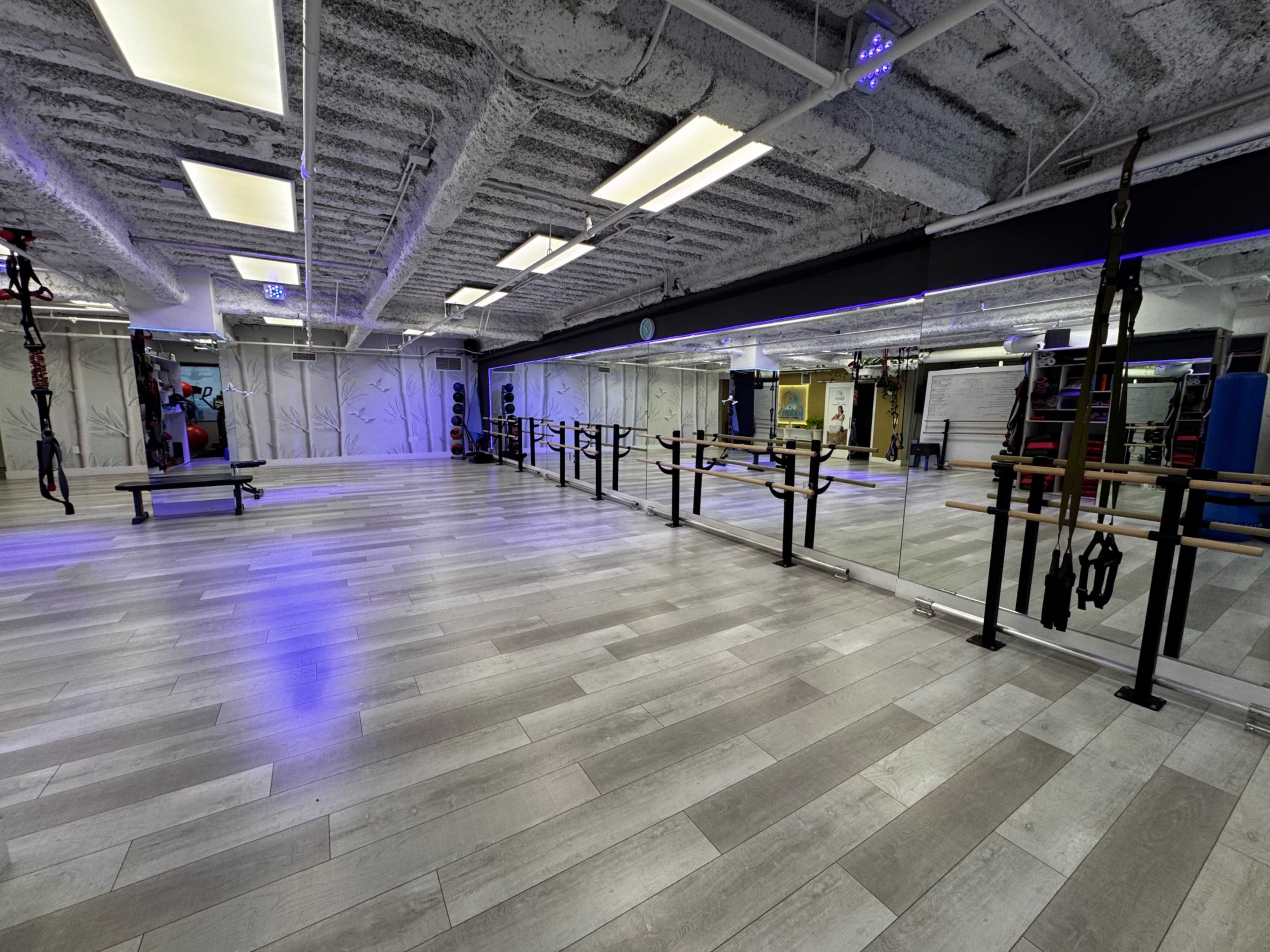 The image shows a bright, spacious fitness studio with wooden floors, mirrored walls, and a row of exercise equipment along one side.