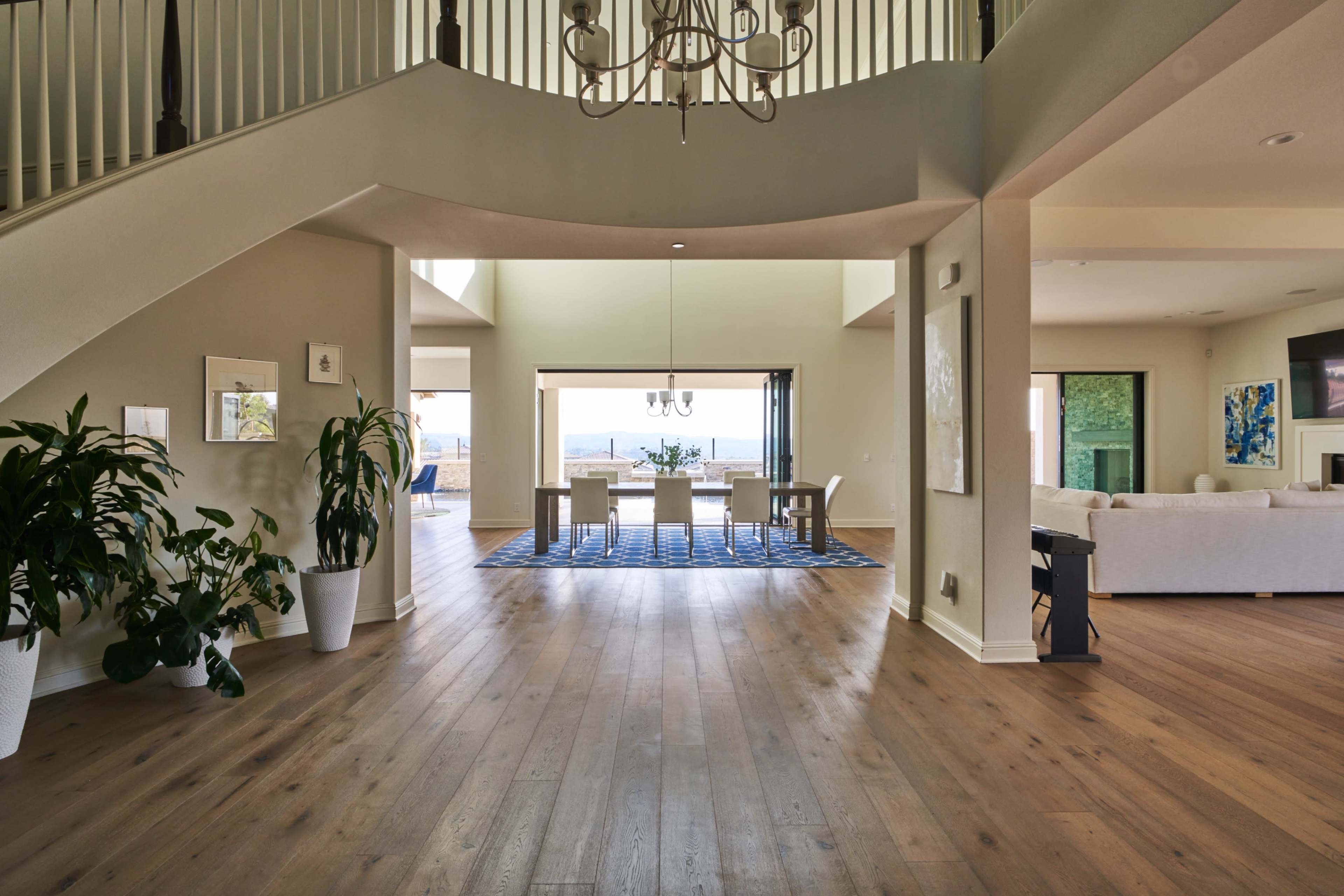 A spacious entryway with a staircase leading to an upper level, opening into a dining area with a table and chairs, visible through large windows.