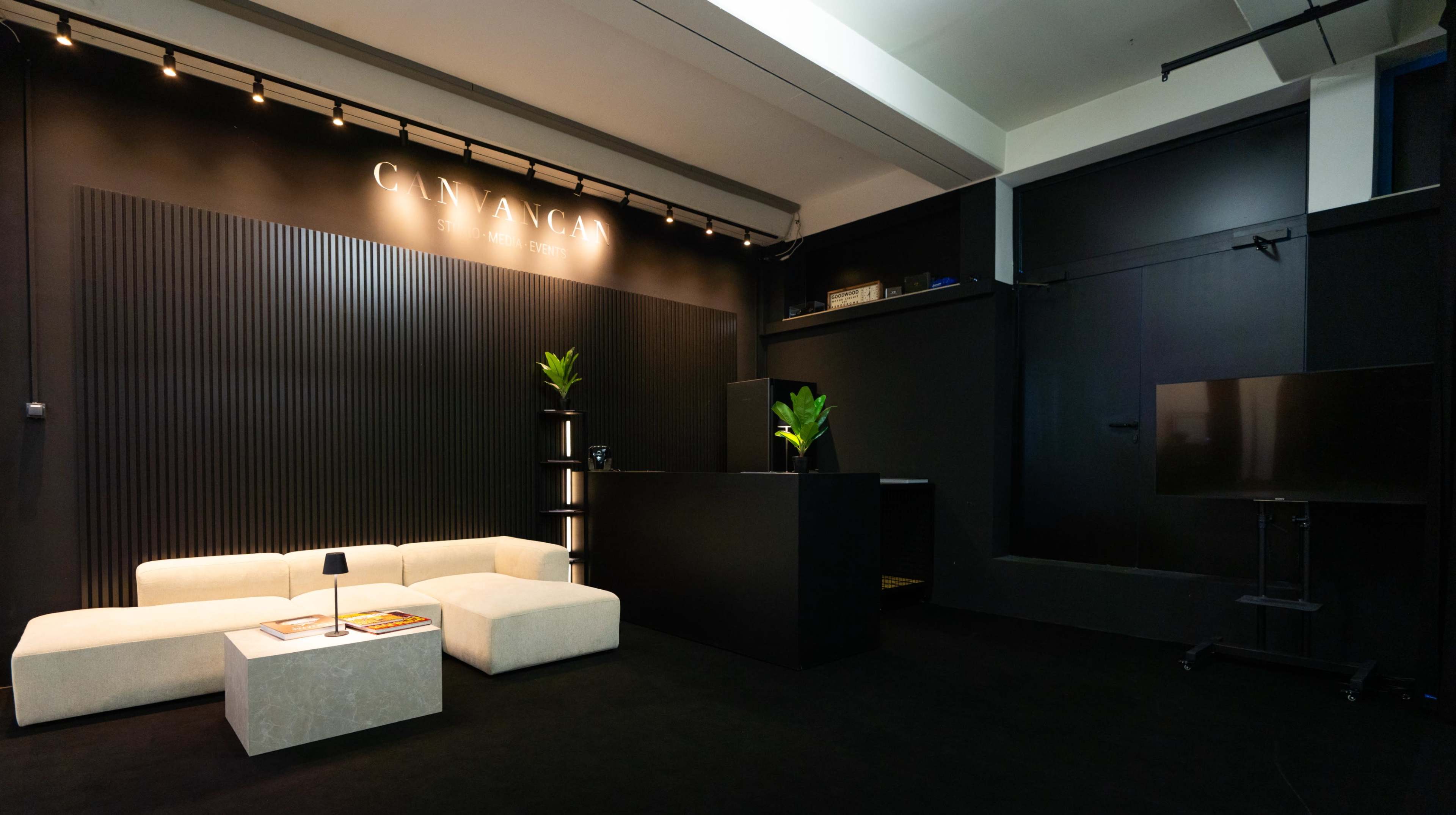The image shows a modern reception area featuring a light-colored sofa, a minimalist black reception desk, and potted plants against a dark wall.