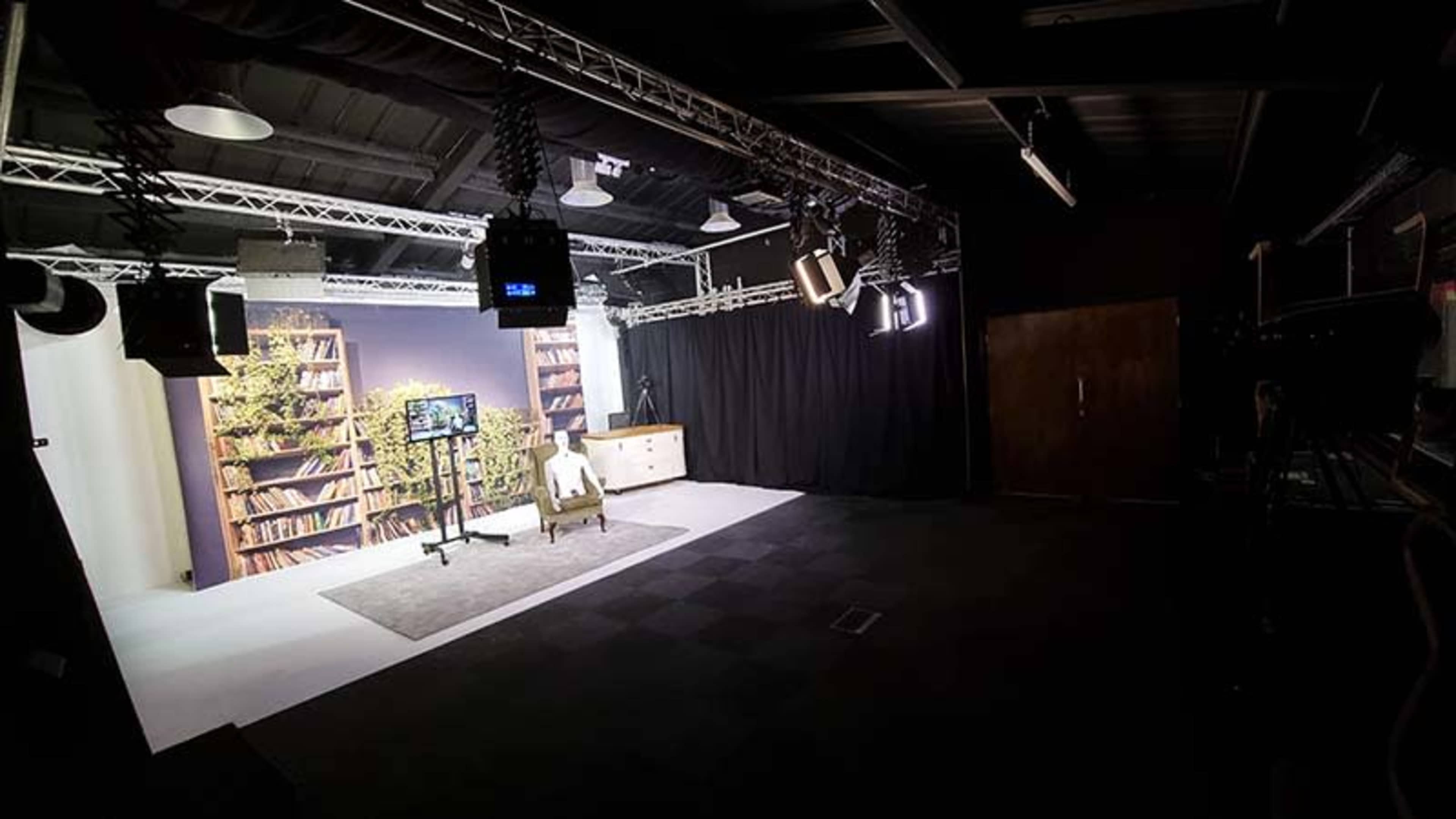 A dimly lit studio space with a single chair facing a backdrop of bookshelves, surrounded by studio lighting and equipment.