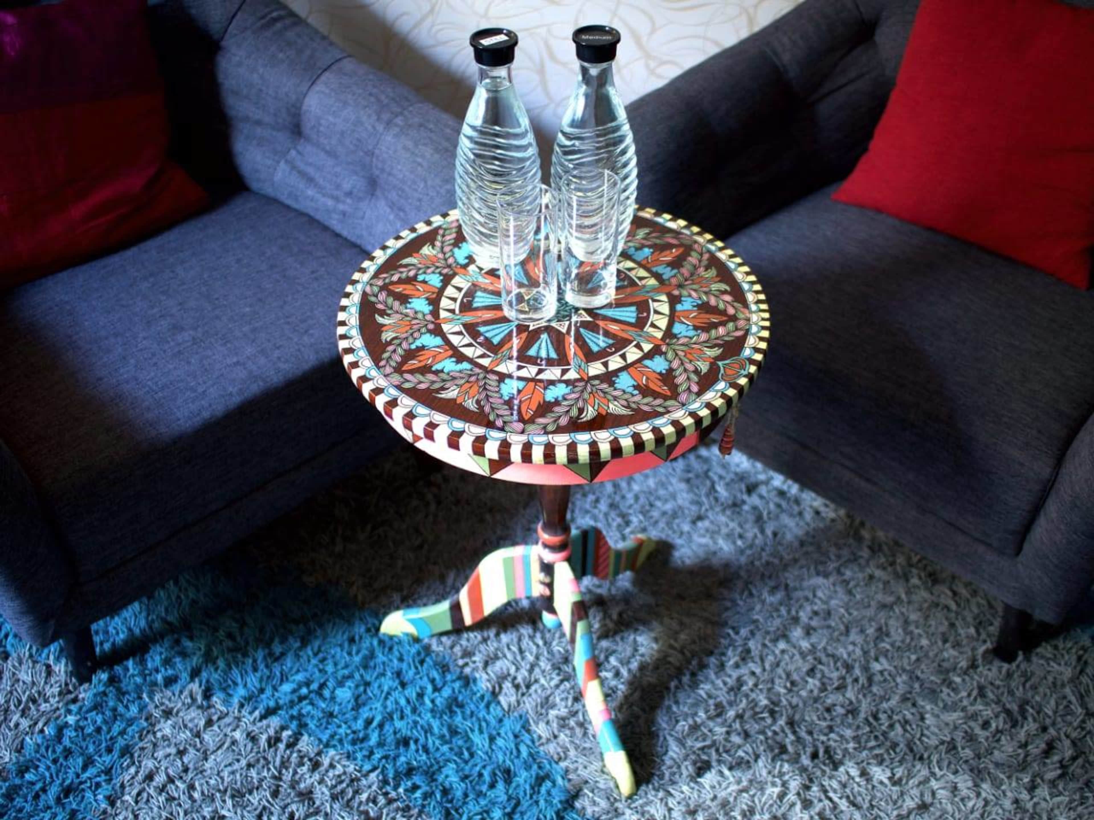 A patterned side table with a colorful design holds two clear water bottles, placed between two gray sofas with red cushions on a textured rug.