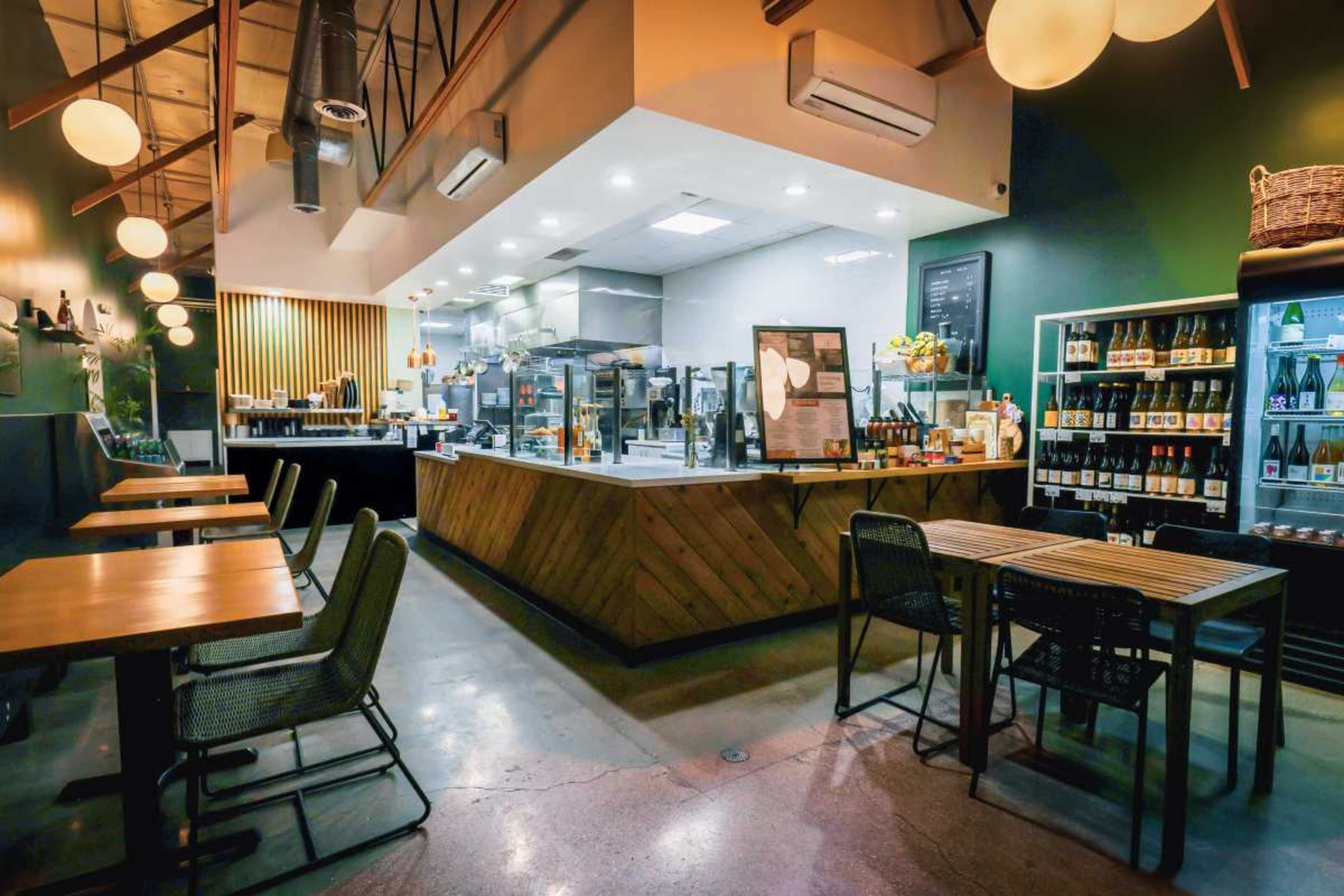 The image shows a modern café with a wooden counter, a well-lit seating area featuring tables and chairs, and a display refrigerator filled with beverages.