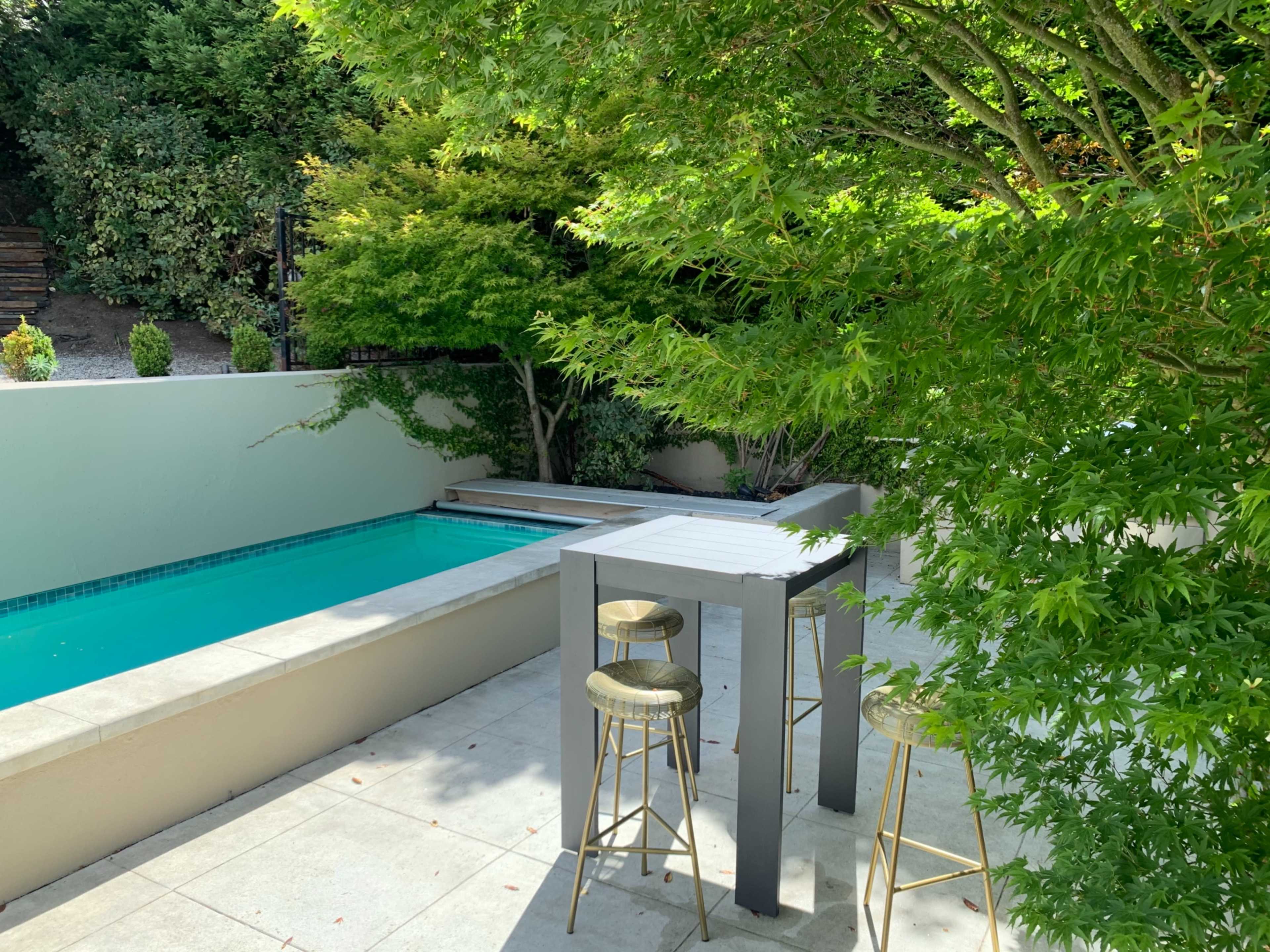 A small outdoor area features a pool bordered by a light-colored wall and a tree, with two high stools positioned near a gray table.